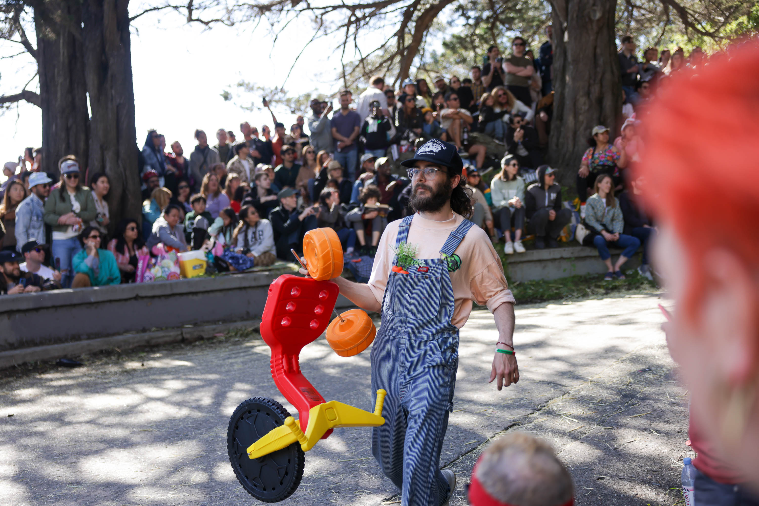 A man in overalls walks with a toy tricycle in front of a crowd outdoors.