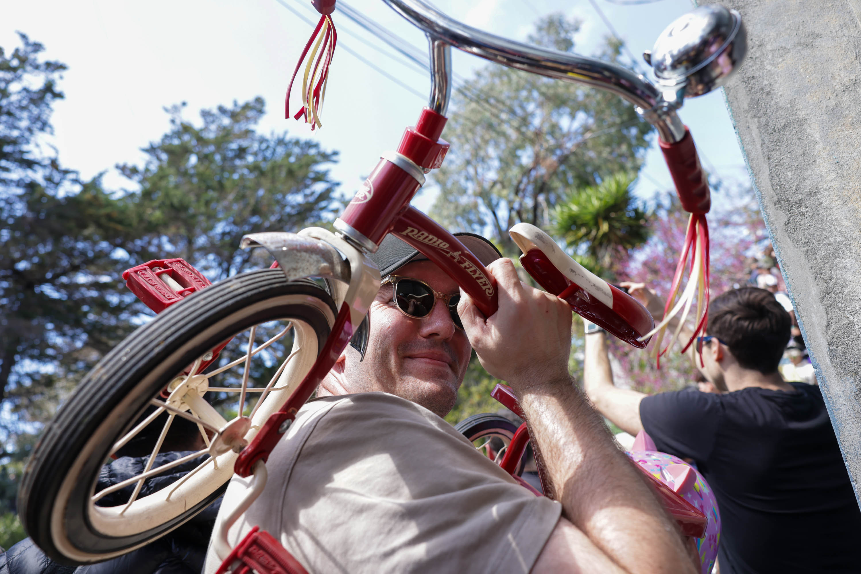 A man peers through the frame of an upside-down red tricycle, its wheel and handlebars prominent.