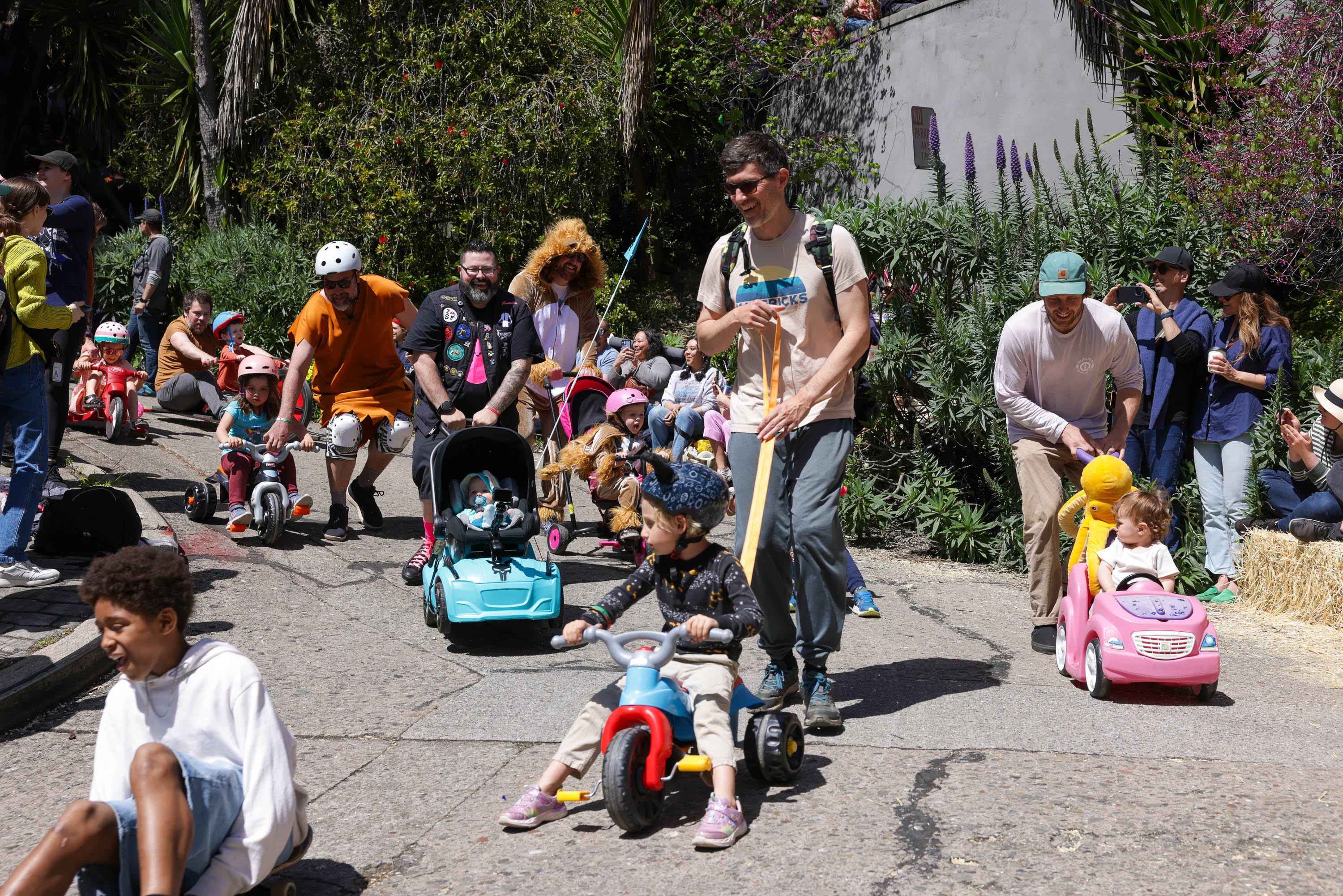A lively outdoor scene where adults and kids happily participate in a toy vehicle parade on a sunny day, with spectators lining the path.