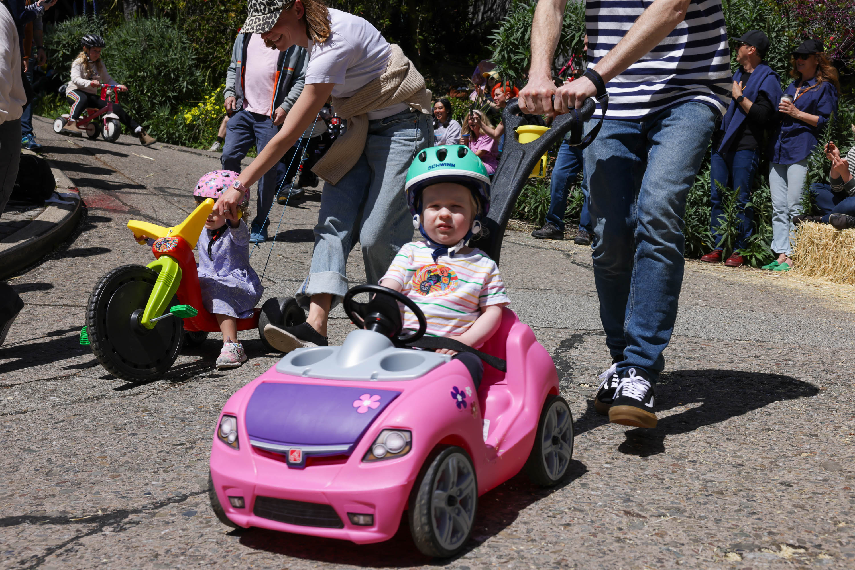 A toddler in a green helmet rides in a pink toy car, pushed by an adult, in a sunny outdoor setting with spectators.