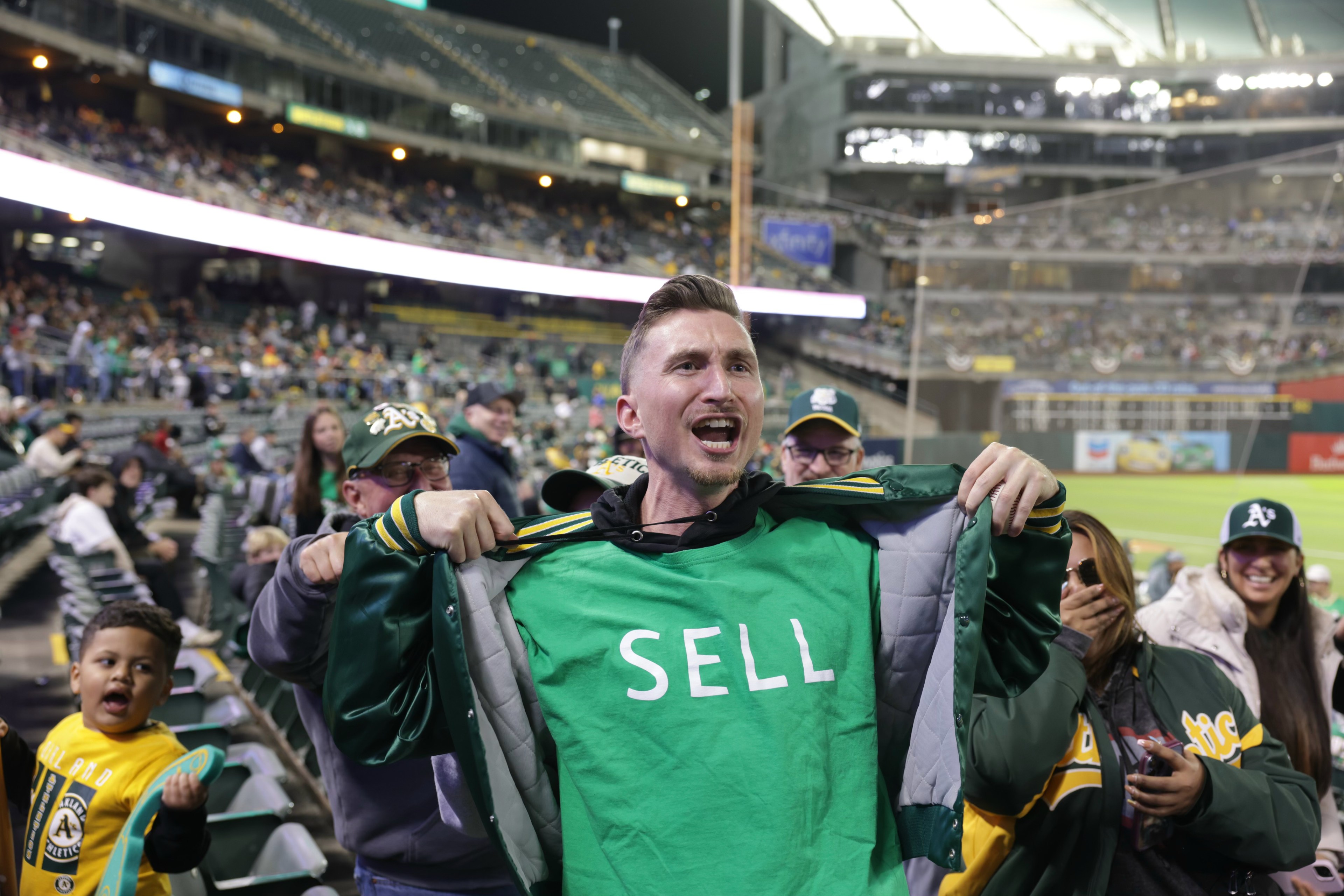 A man at a sports stadium is holding open his jacket to reveal a green shirt with the word &quot;SELL&quot; printed on it. Other people around him are smiling and cheering.