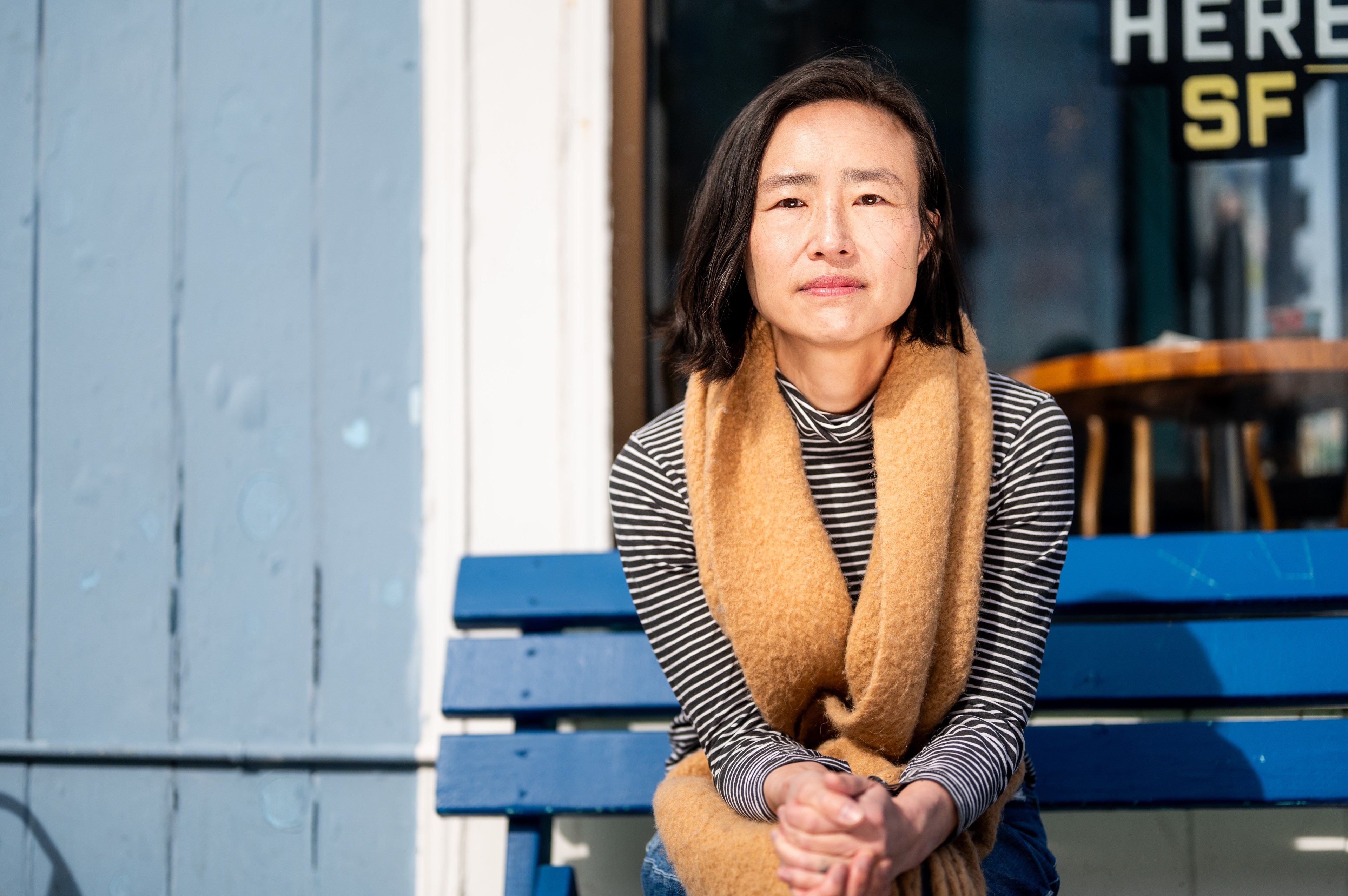 A woman with a short black bob hairstyle is sitting on a blue bench. She wears a striped shirt with a brown scarf and is illuminated by the sun against a blue wall.