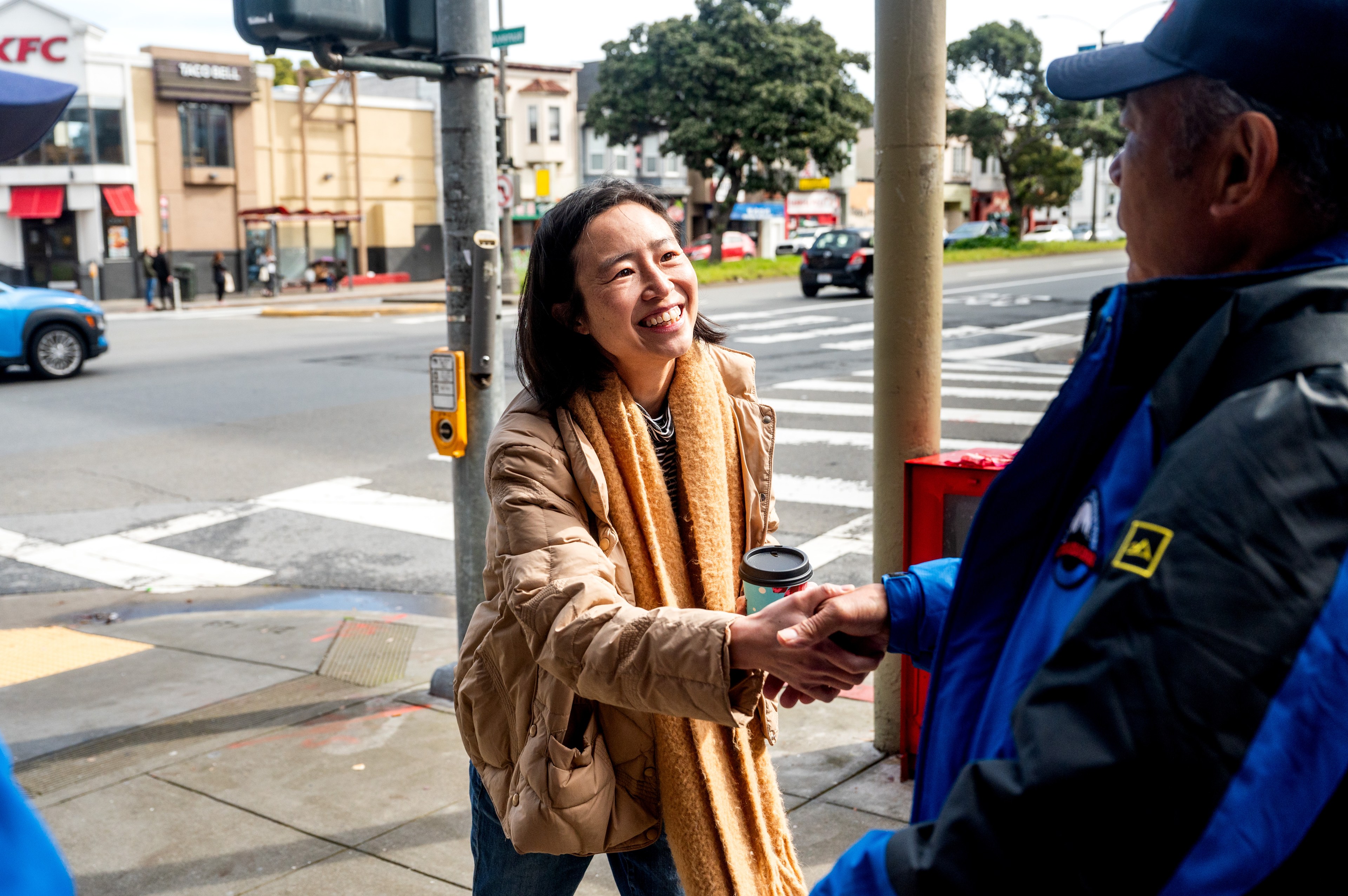 A smiling woman shaking hands with a man on a city street corner, both holding coffee cups.