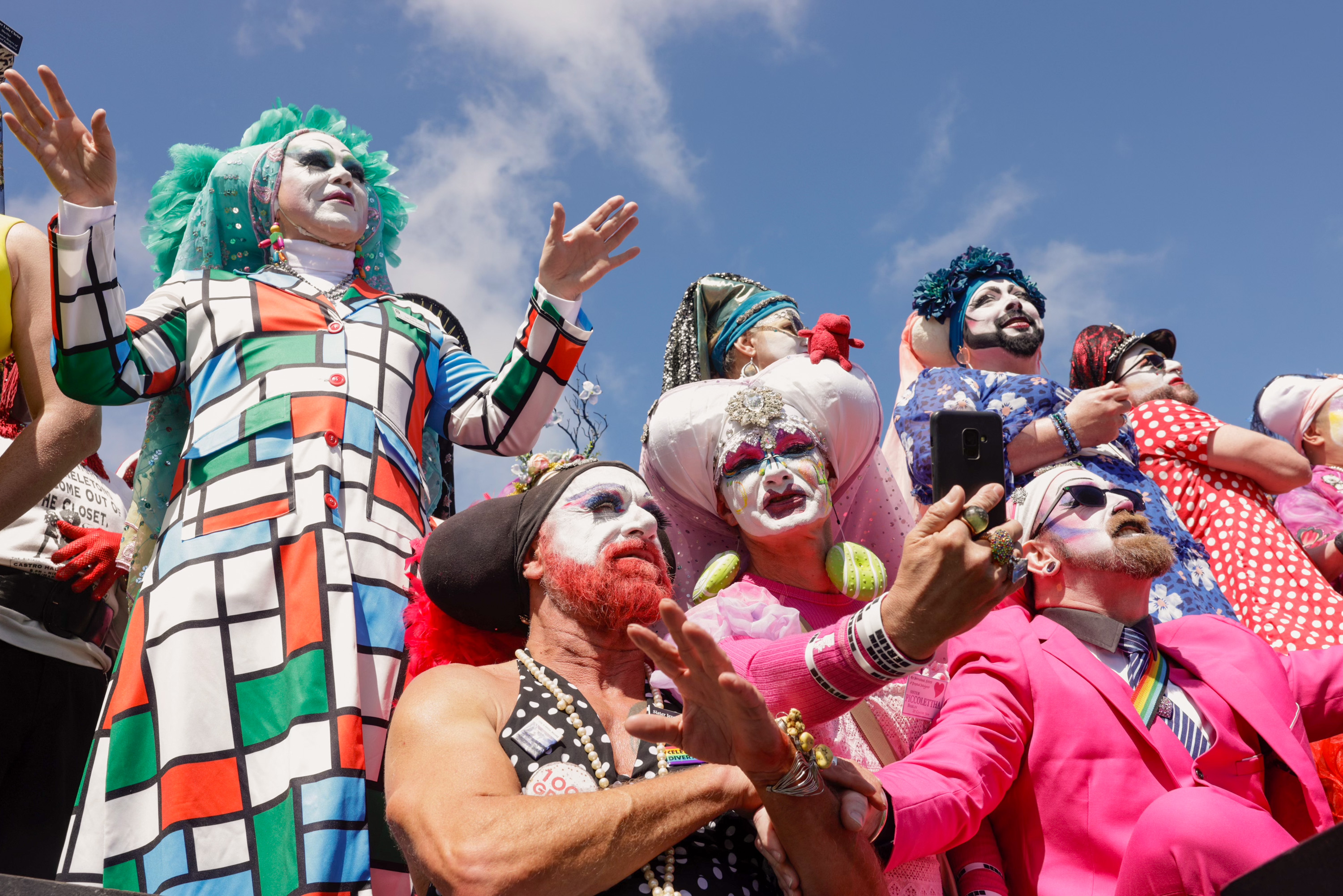 A group of people in colorful, exaggerated costumes and makeup, likely at a festive event, posing with theatrical expressions under a blue sky.