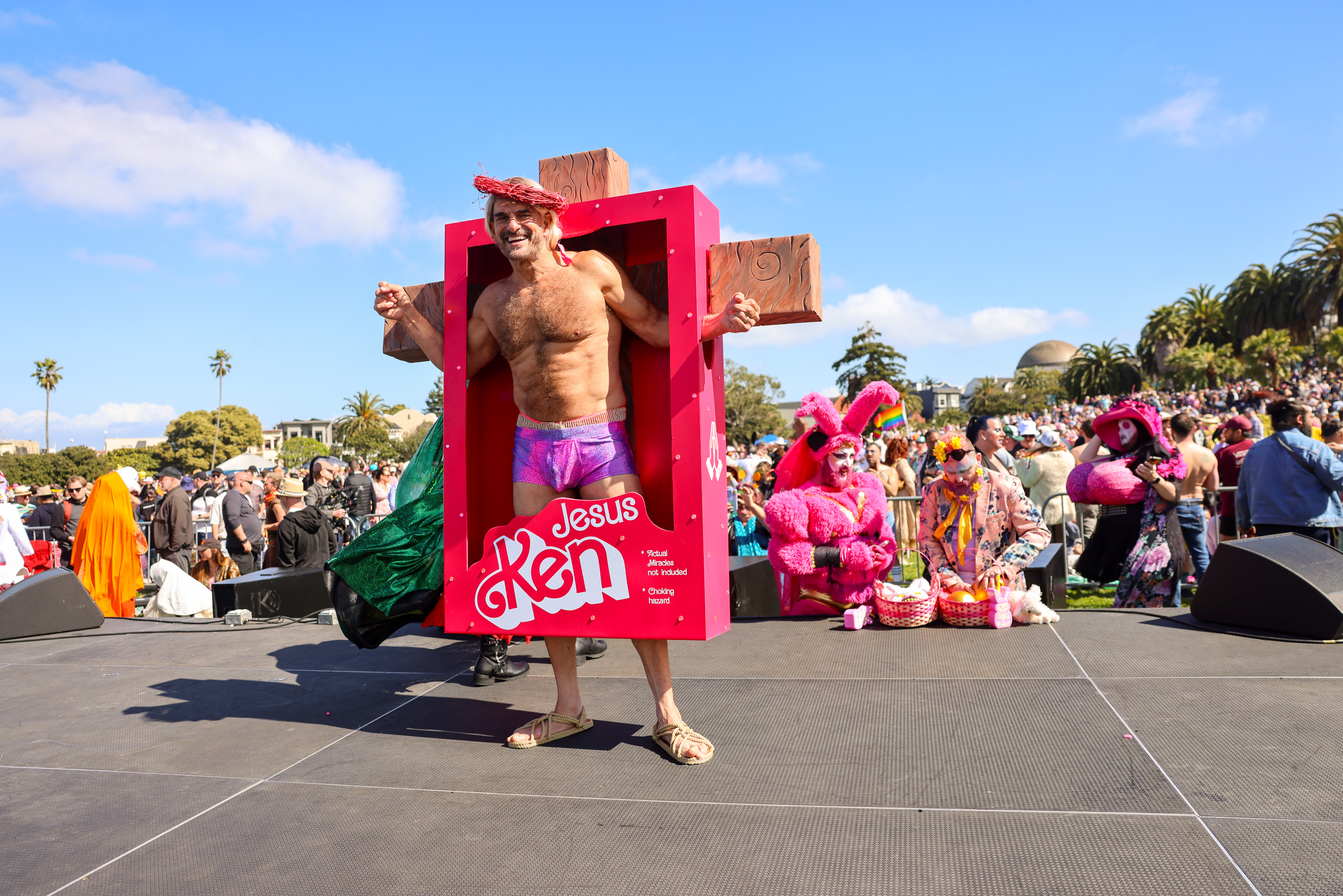 A man in pink shorts pops out of a box labeled &quot;Jesus Ken,&quot; on stage at a festive event with costumed people around.