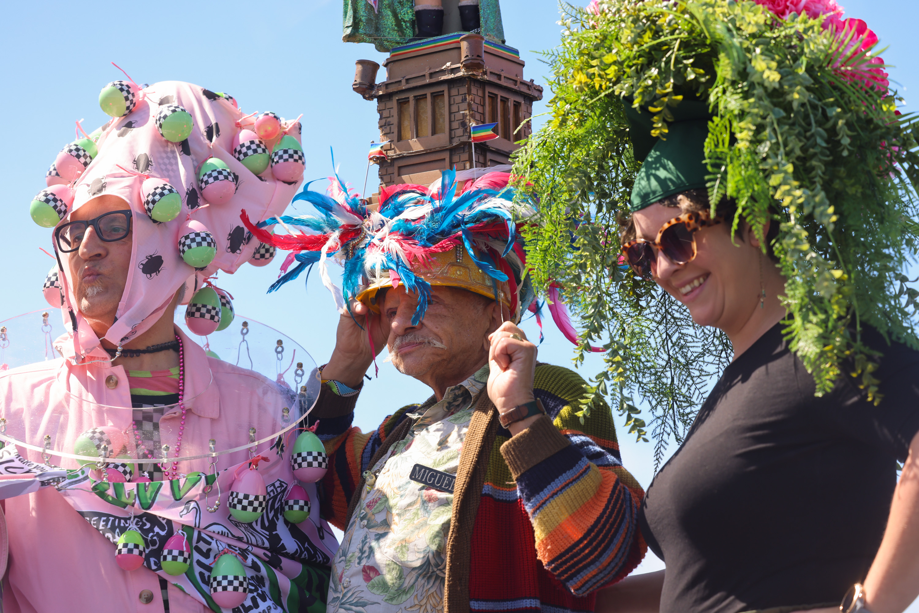 Three people in whimsical costumes with colorful hats, one resembling a flower arrangement, at a bright outdoor event.