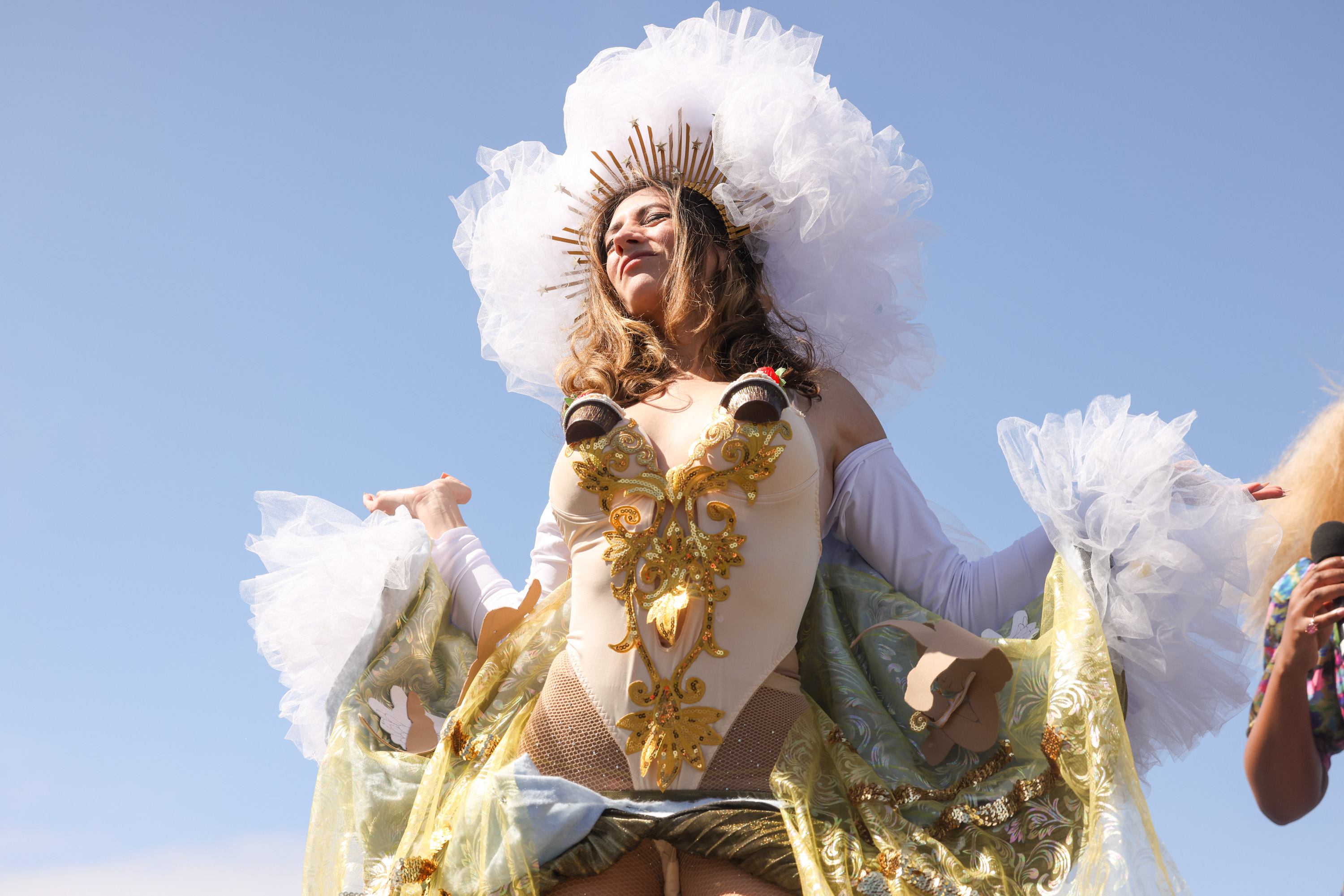 A person in a lavish costume with a large white headpiece is at an event. They exude confidence under a clear sky.