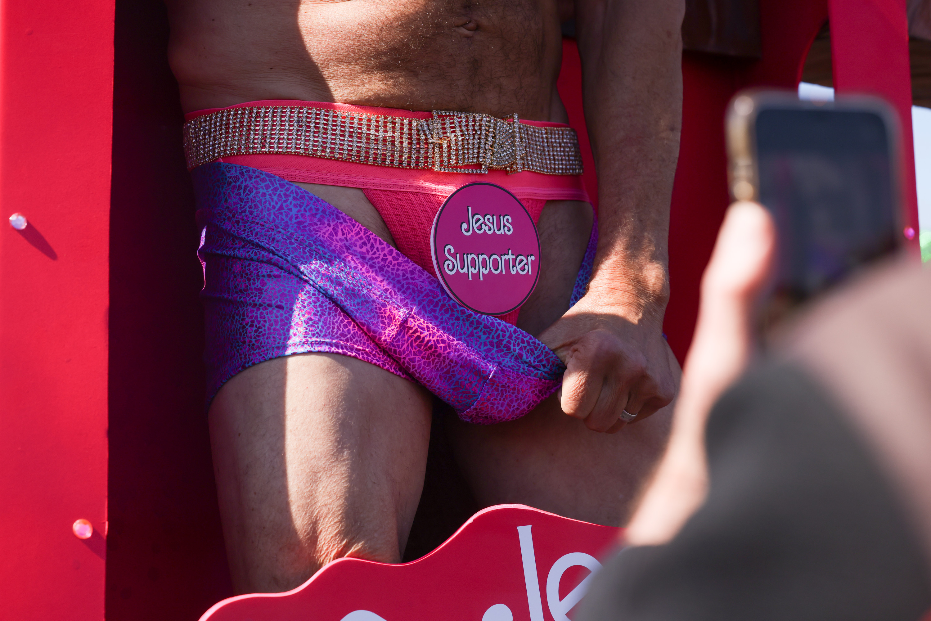 A person is wearing a pink, bedazzled belt with a &quot;Jesus Supporter&quot; badge and a purple garment. A smartphone is capturing the scene.