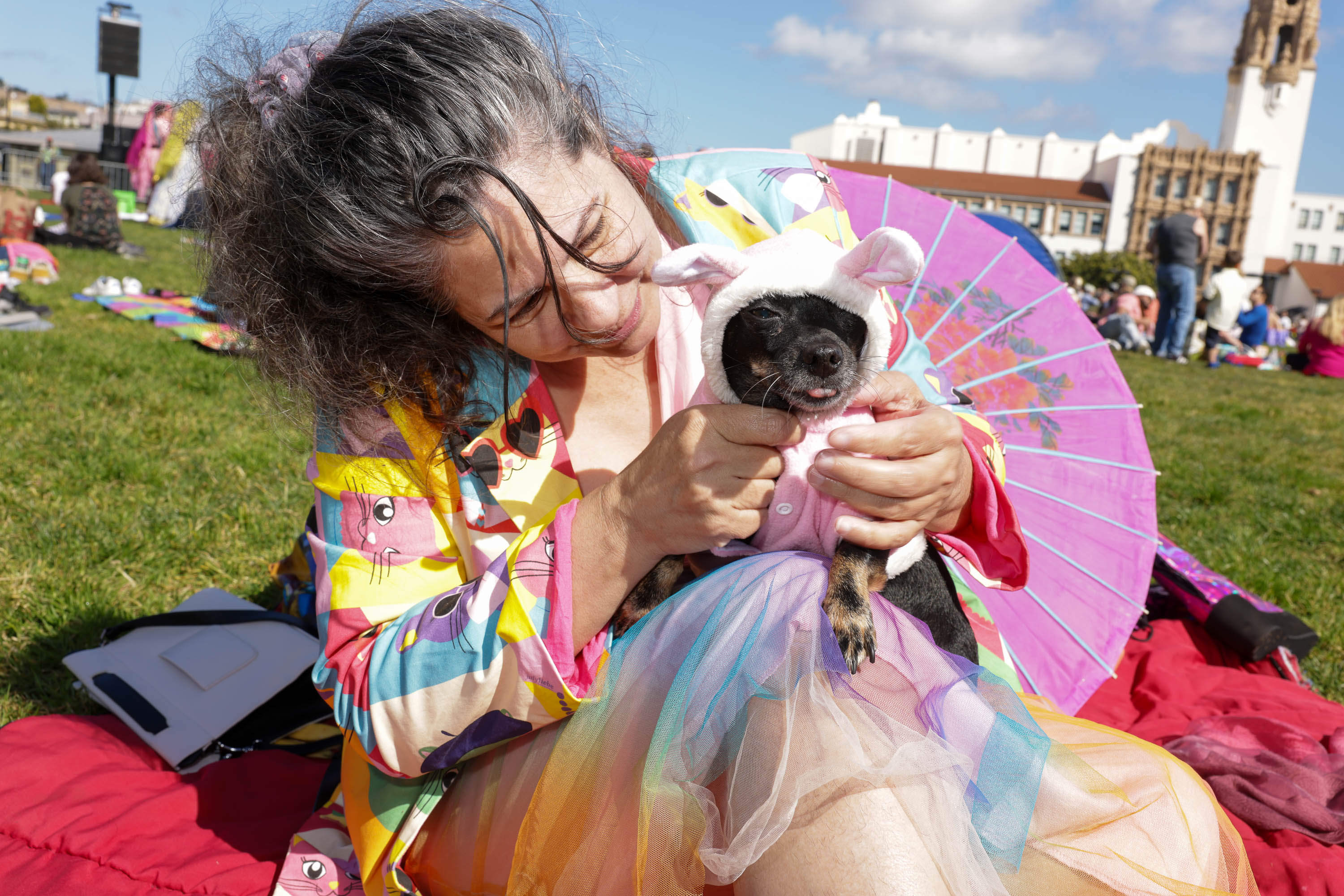 A woman in colorful attire cuddles a dog in a pink outfit and hat, with a pink fan behind them, outdoors on a sunny day.