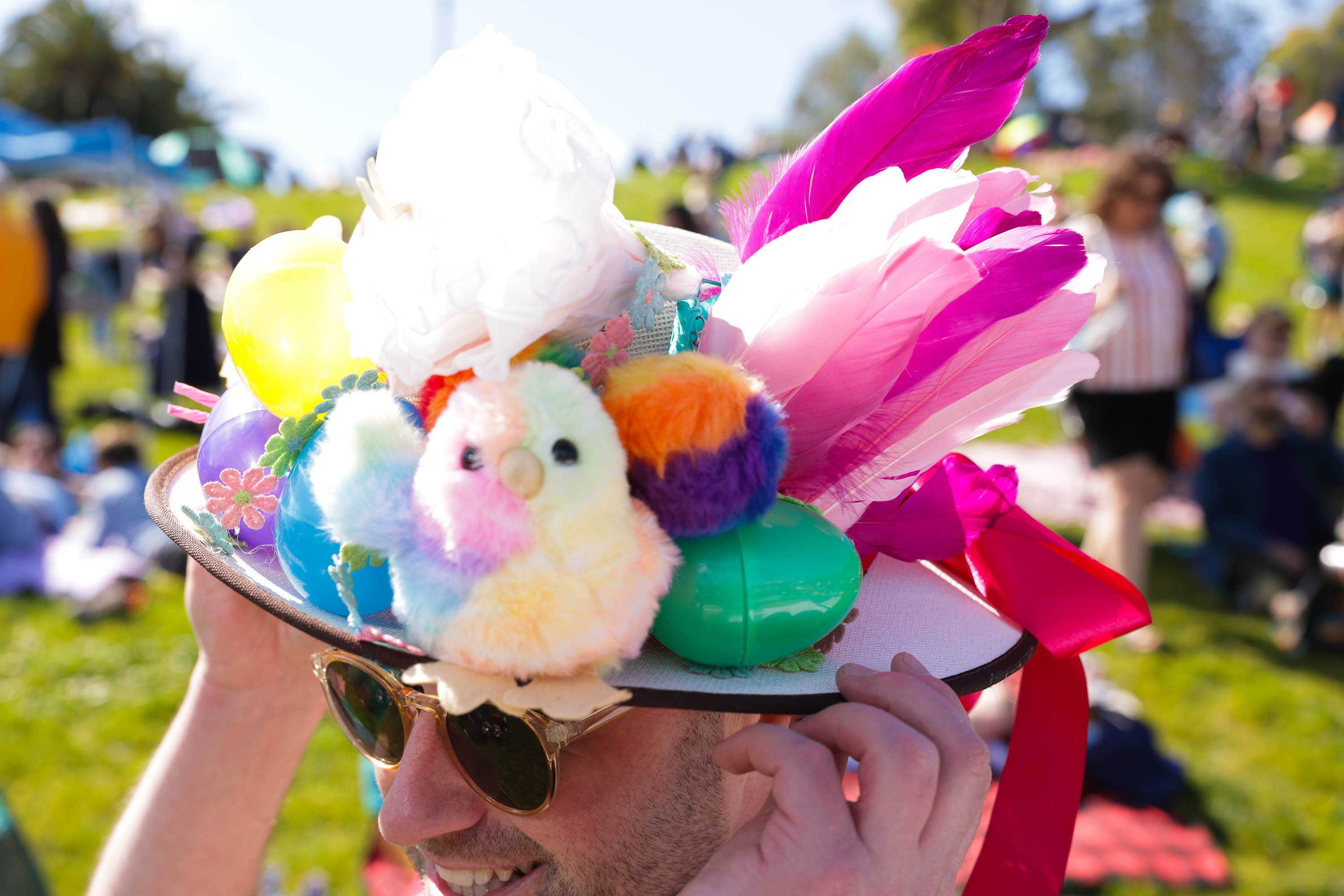 A person wearing a hat adorned with colorful feathers, balloons, and a plush toy, at an outdoor event.