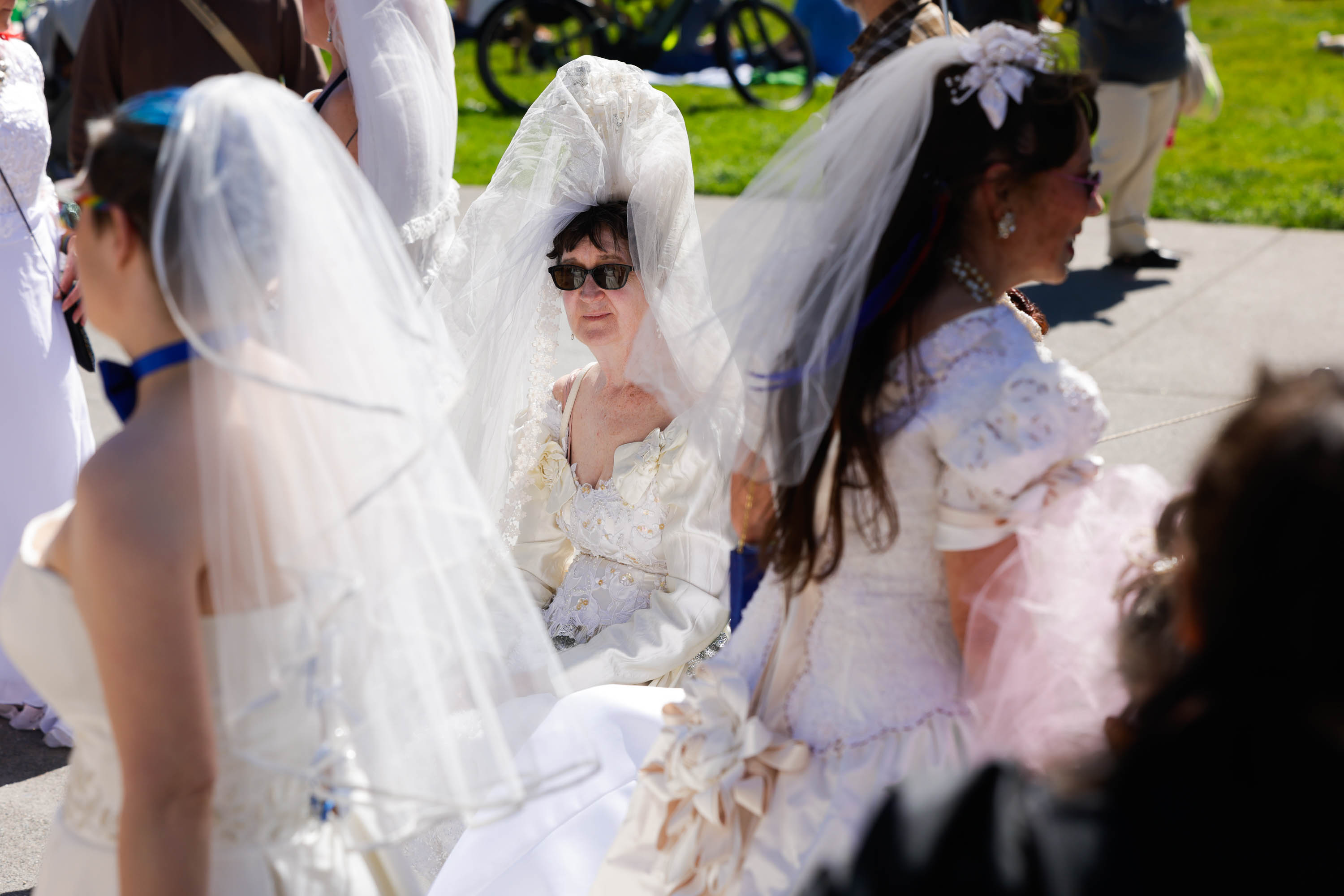 A group of people wearing various wedding dresses and veils gather outdoors.
