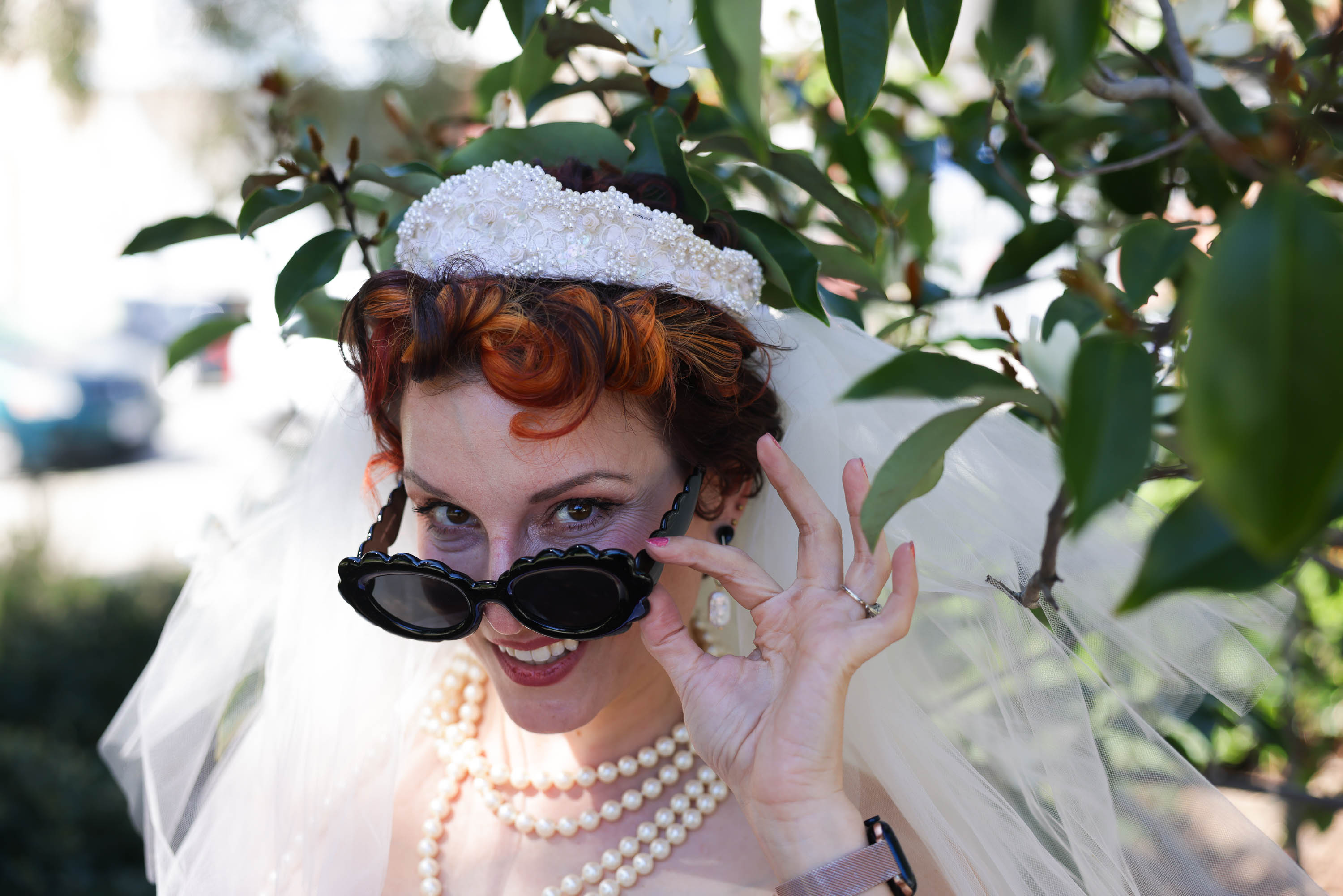 A bride with red hair peeks over her sunglasses, smiling playfully amidst green foliage.
