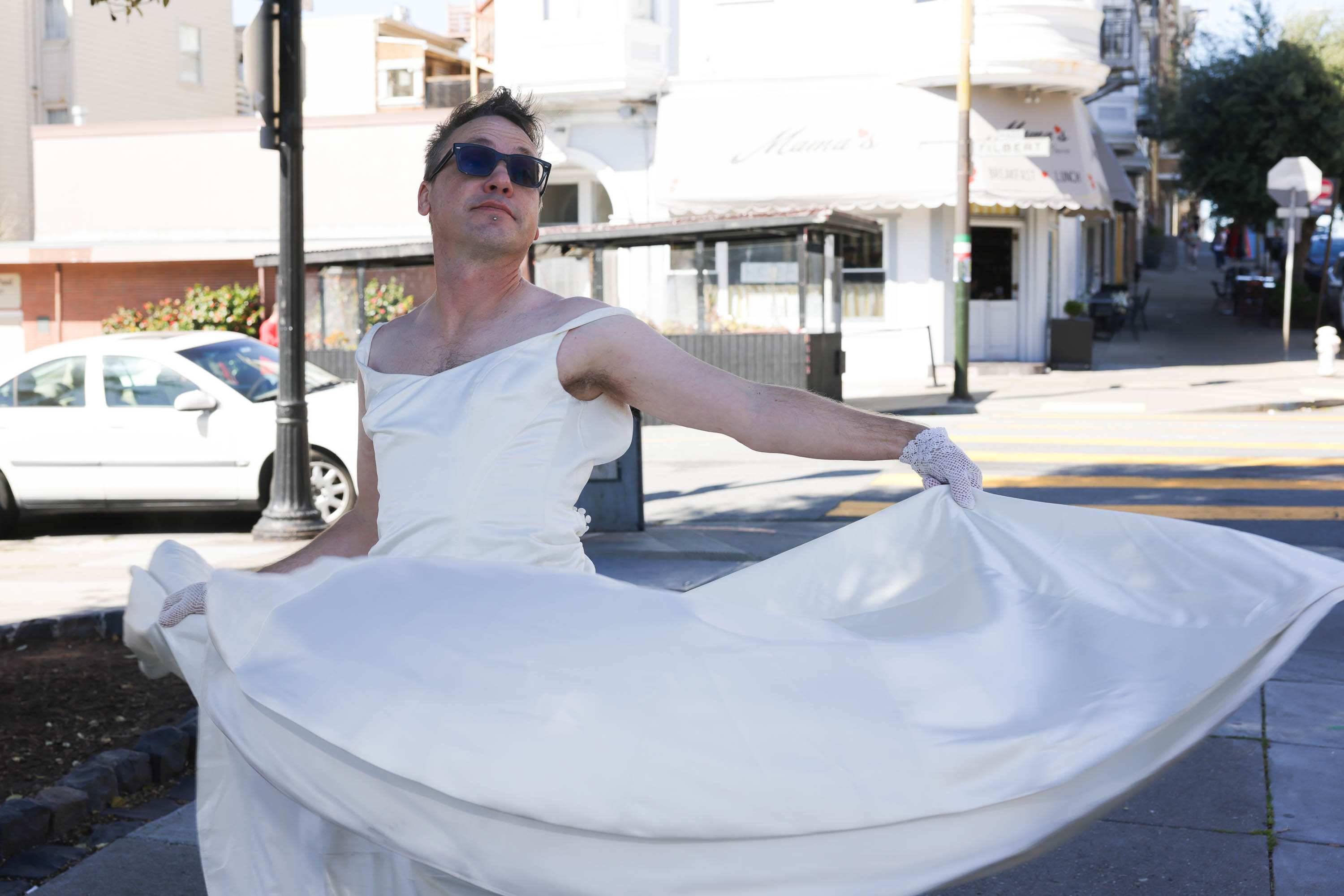 A man in sunglasses poses in a white dress, on a street, with a quirky expression.