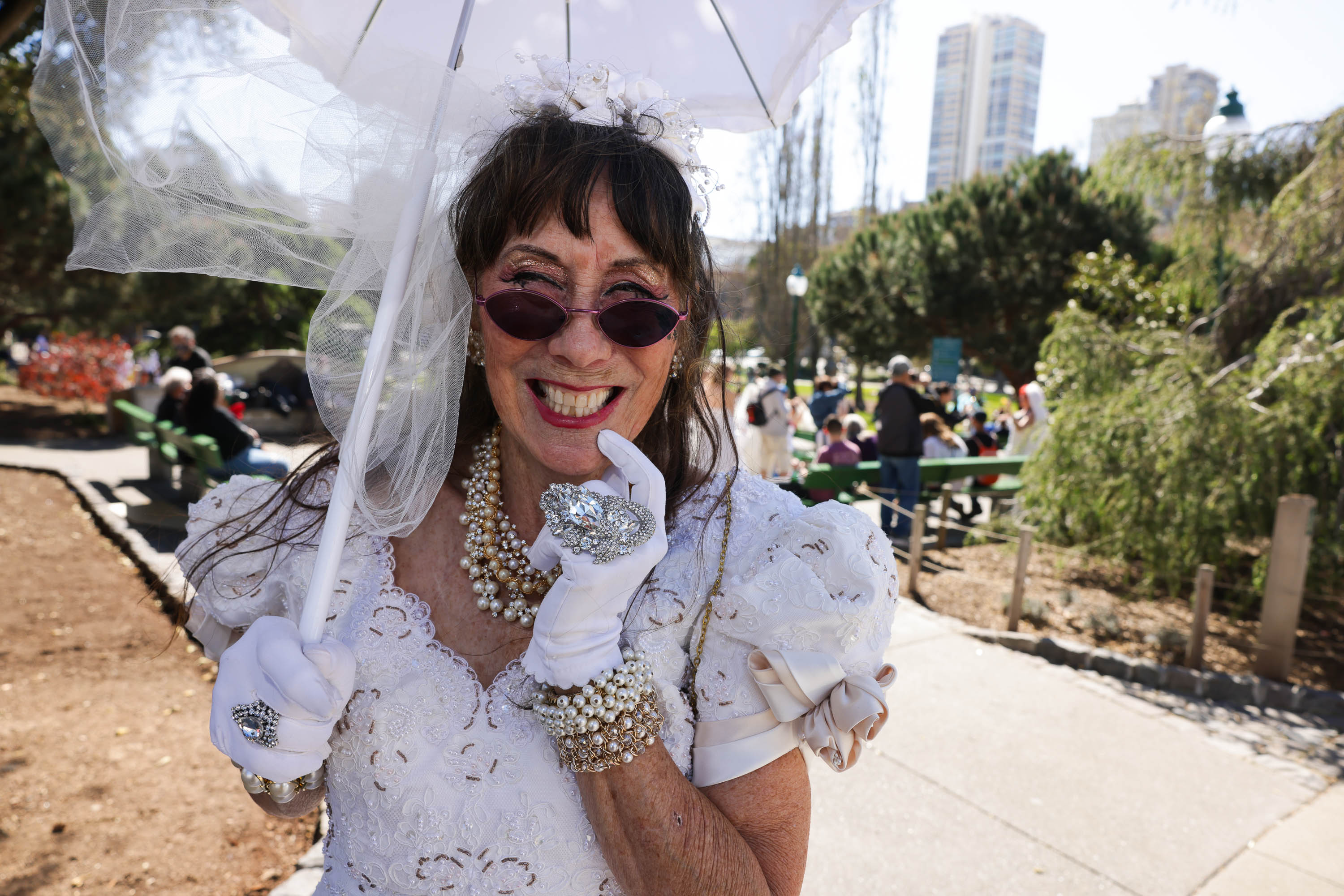 A smiling woman in a white bridal outfit wears sunglasses and pearls and holds an umbrella in an outdoor setting with people in the background.