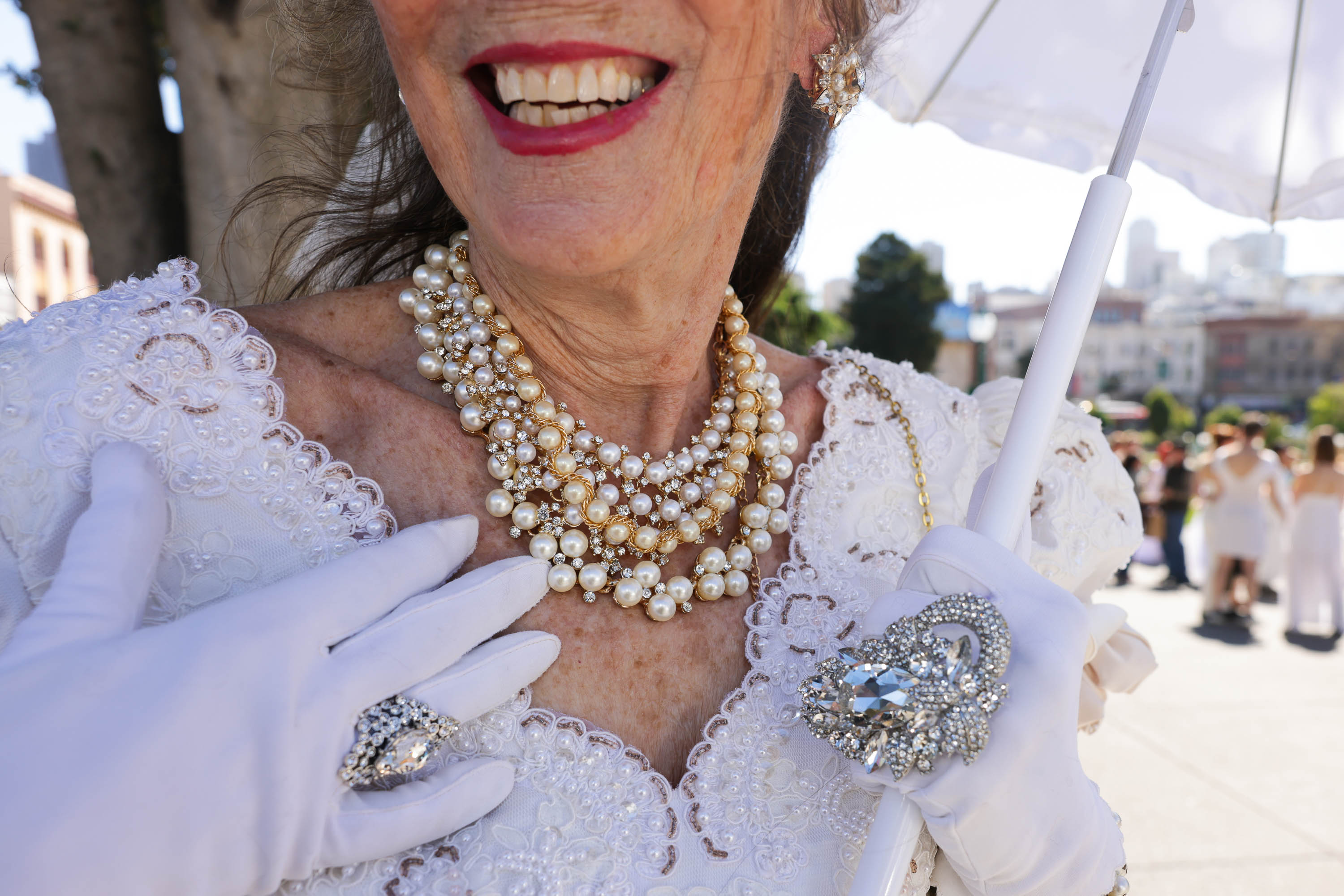 An elegantly dressed older woman smiles, displays a pearl necklace, wears white gloves, and holds a parasol.