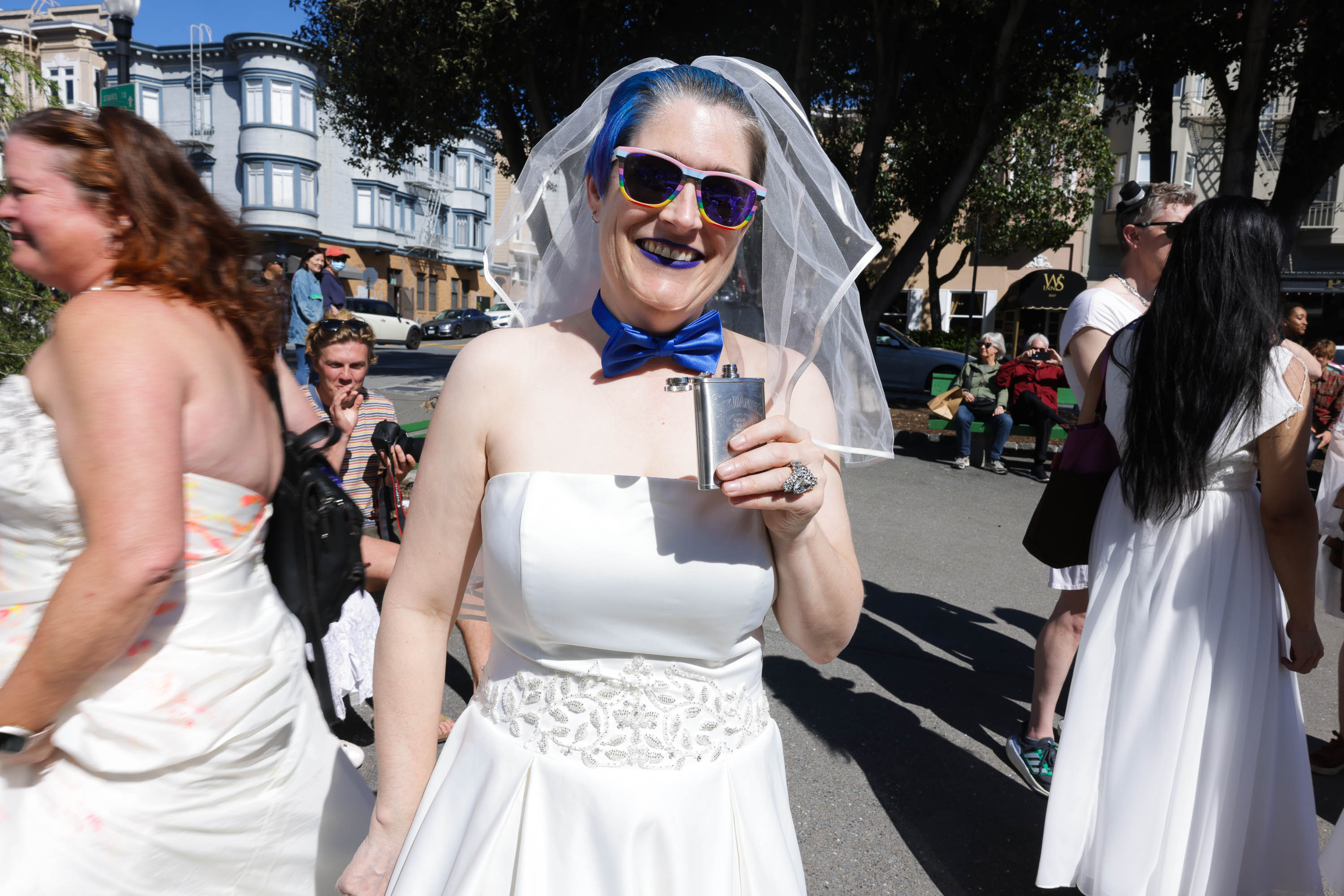 A smiling person in a white dress and veil with blue hair and bowtie holds a flask at a sunny outdoor event.