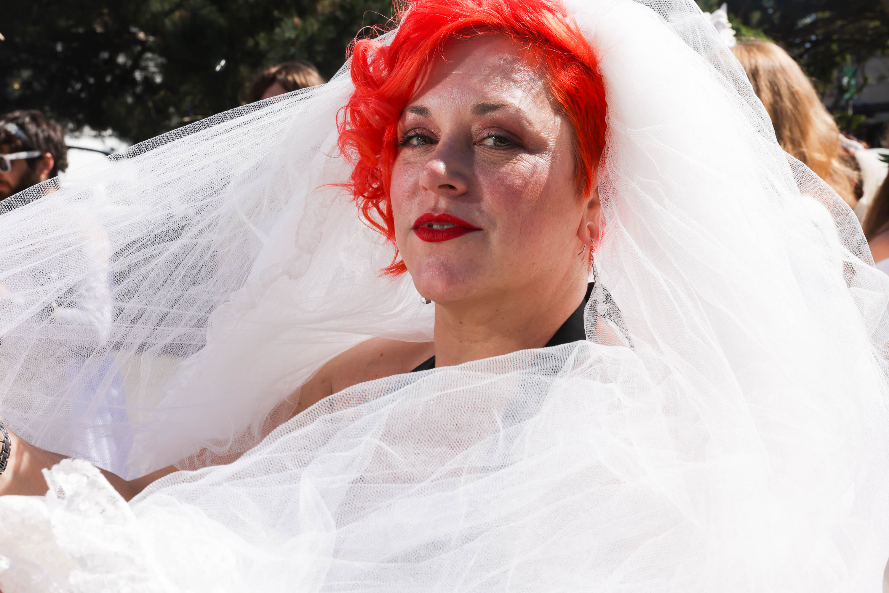 A woman with vibrant red hair is wrapped in tulle veil and wears bright red lipstick and an introspective look.