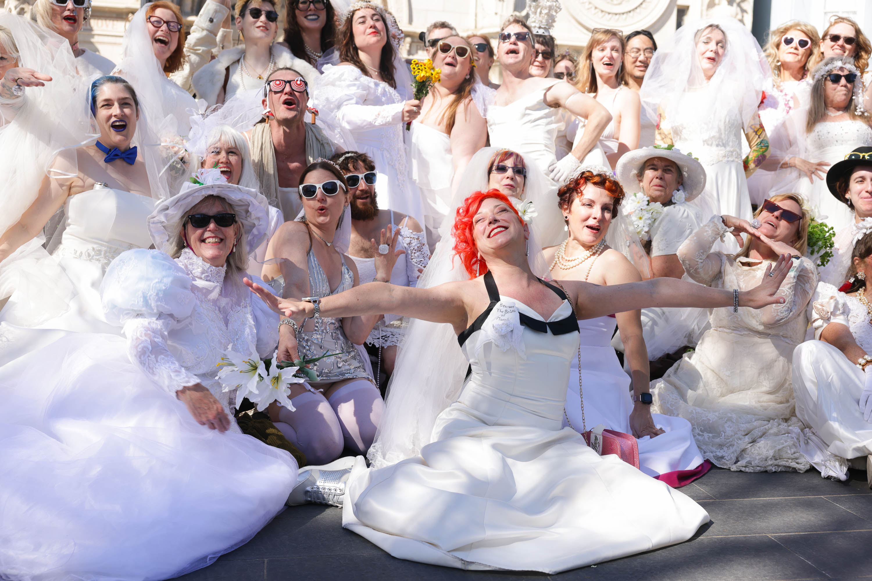 A group of joyful people, some in sunglasses, dressed in various wedding gowns, poses with expressive excitement.