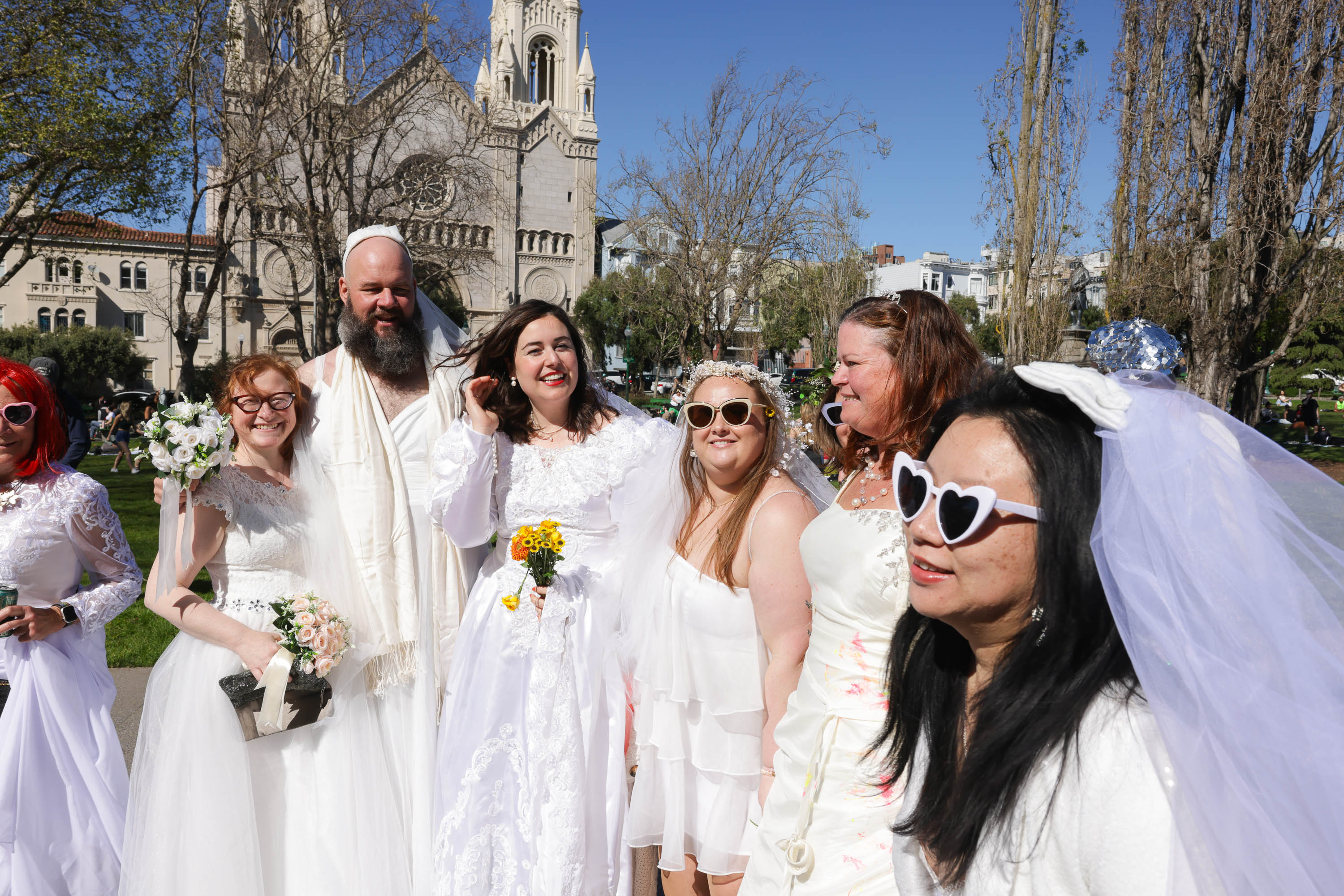 A group of people in wedding attire, smile outdoors, with a historic church in the background.