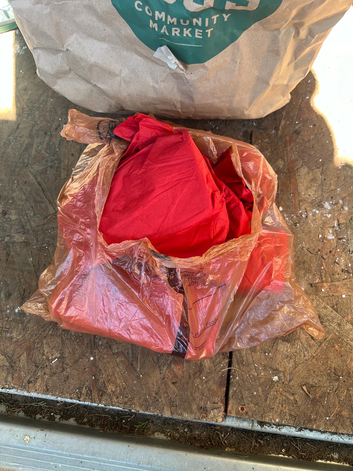 A red cloth is partially visible within a red plastic bag, resting on a wooden surface next to a torn paper bag labeled &quot;COMMUNITY MARKET.&quot;