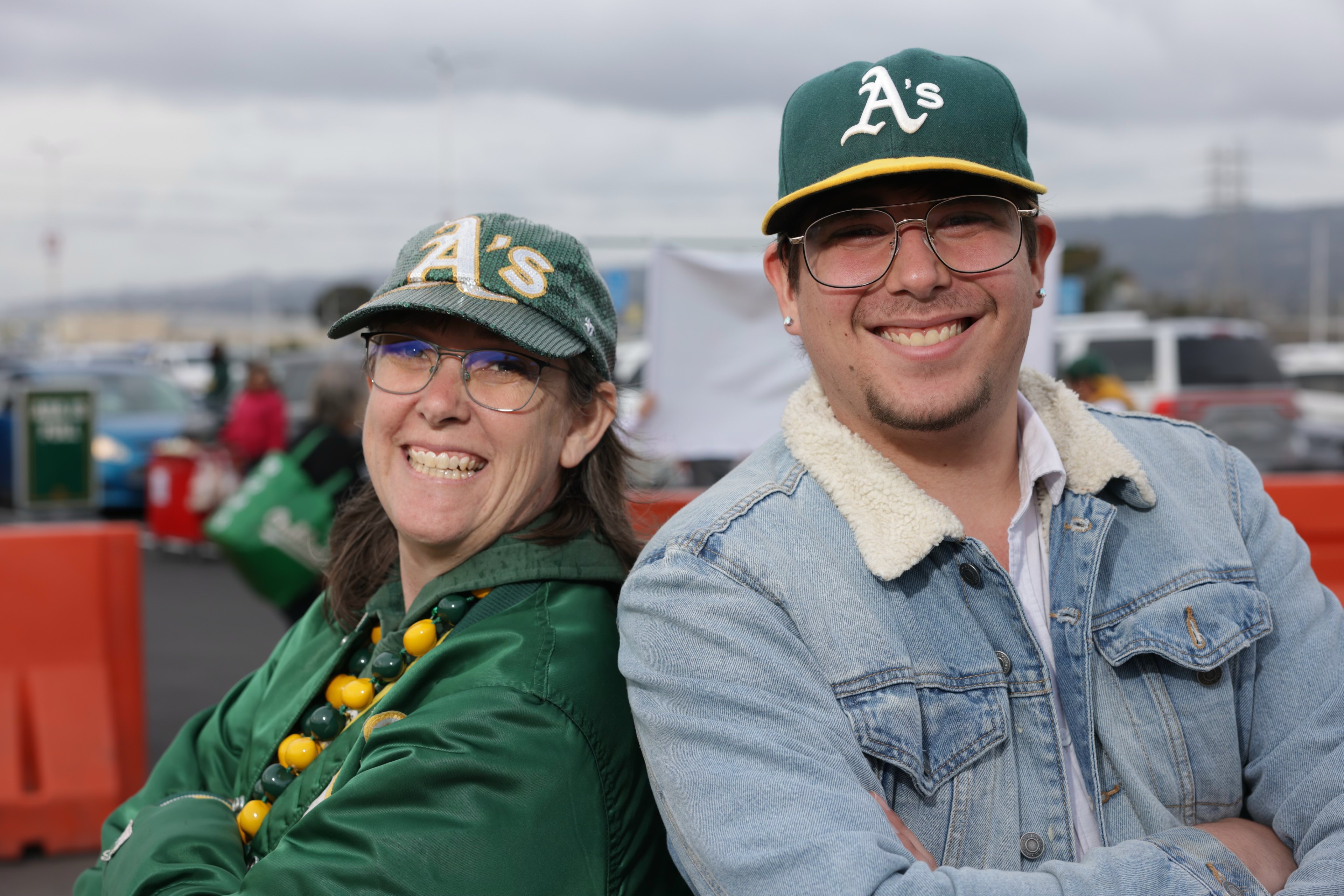 Two people smiling, wearing Oakland A's caps, with a stadium in the background.