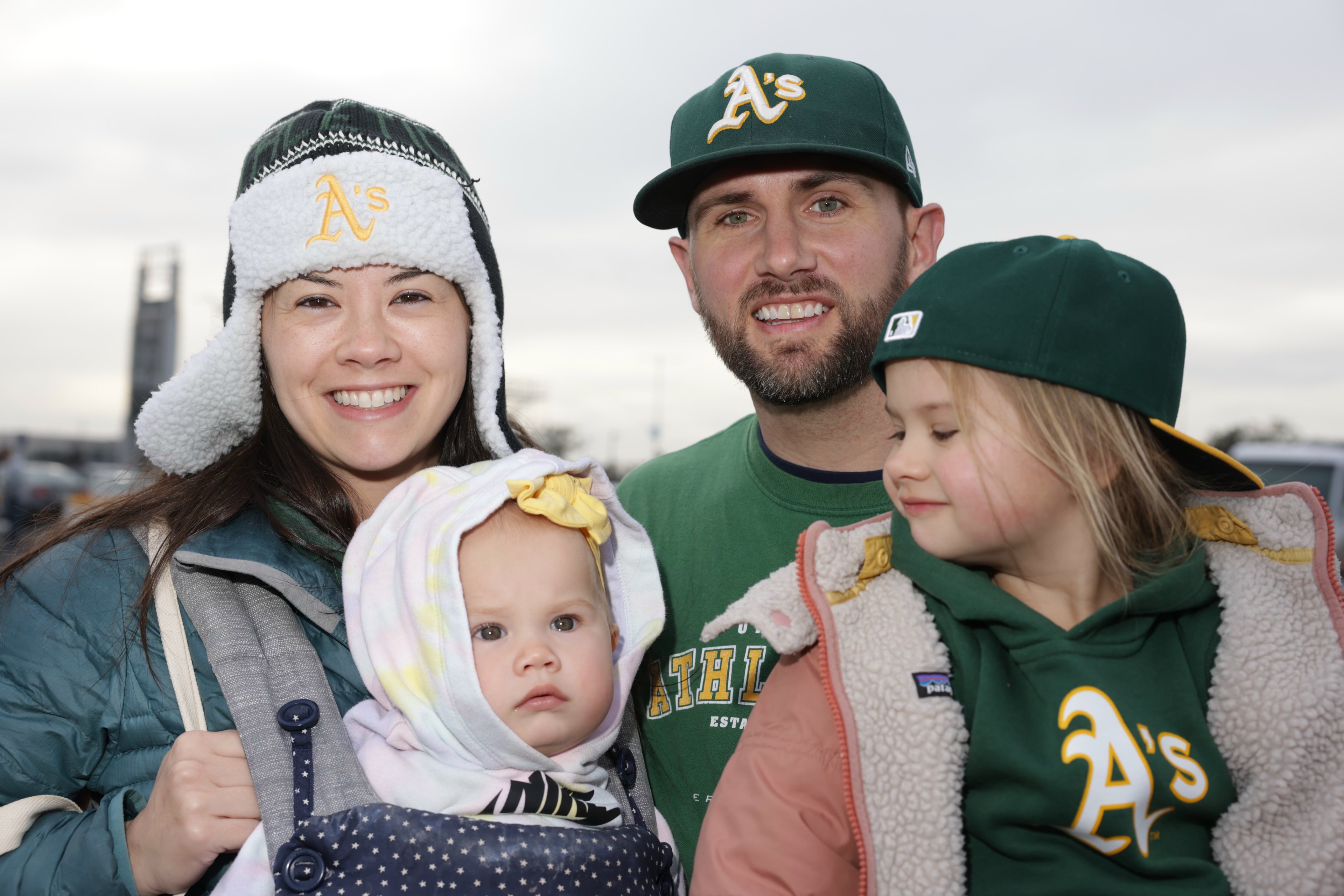 A family of four, clad in Oakland A's gear, smiles outdoors, with two young children in tow.