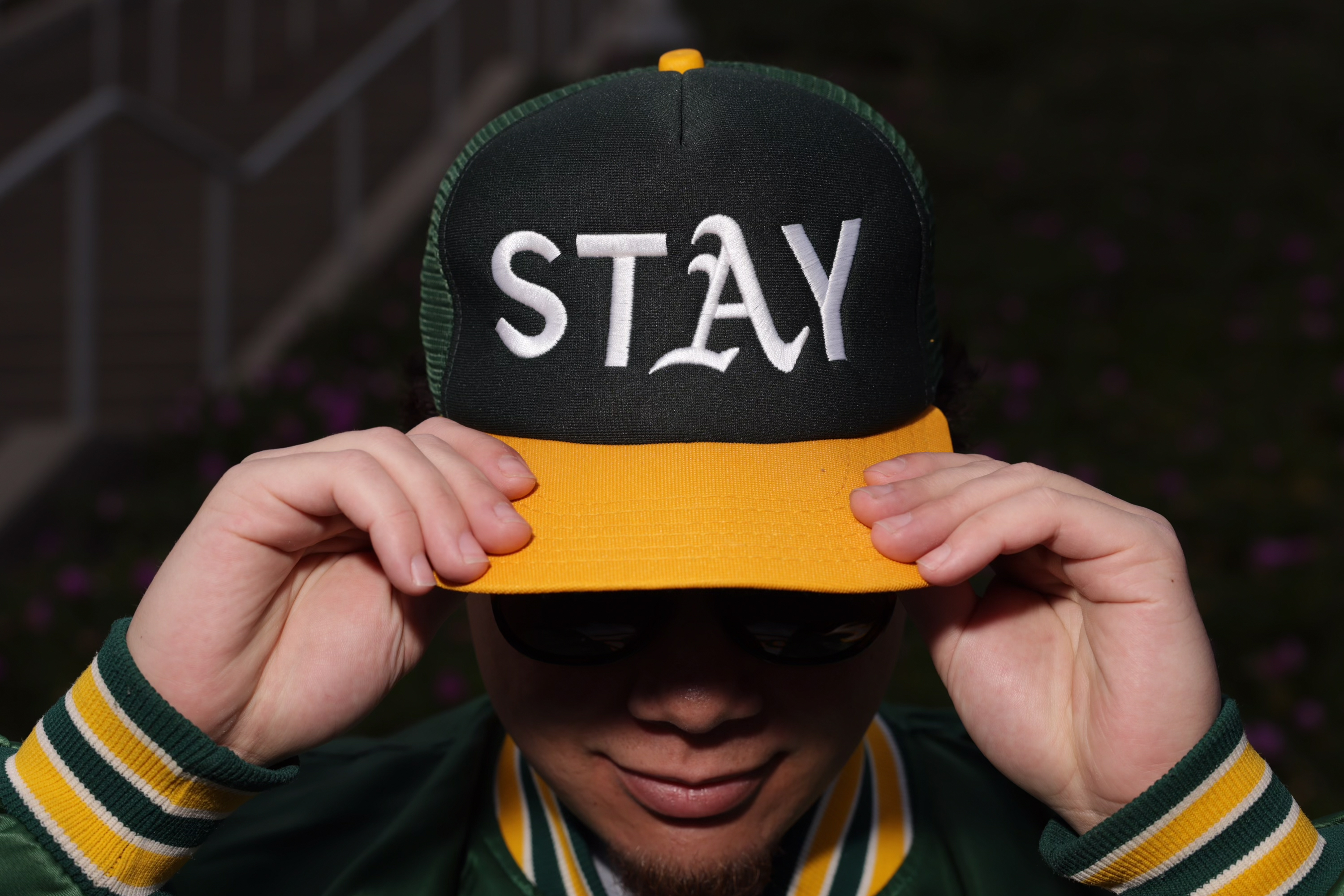 A person in sunglasses adjusts a green and yellow cap with the word &quot;STAY&quot; on the front.