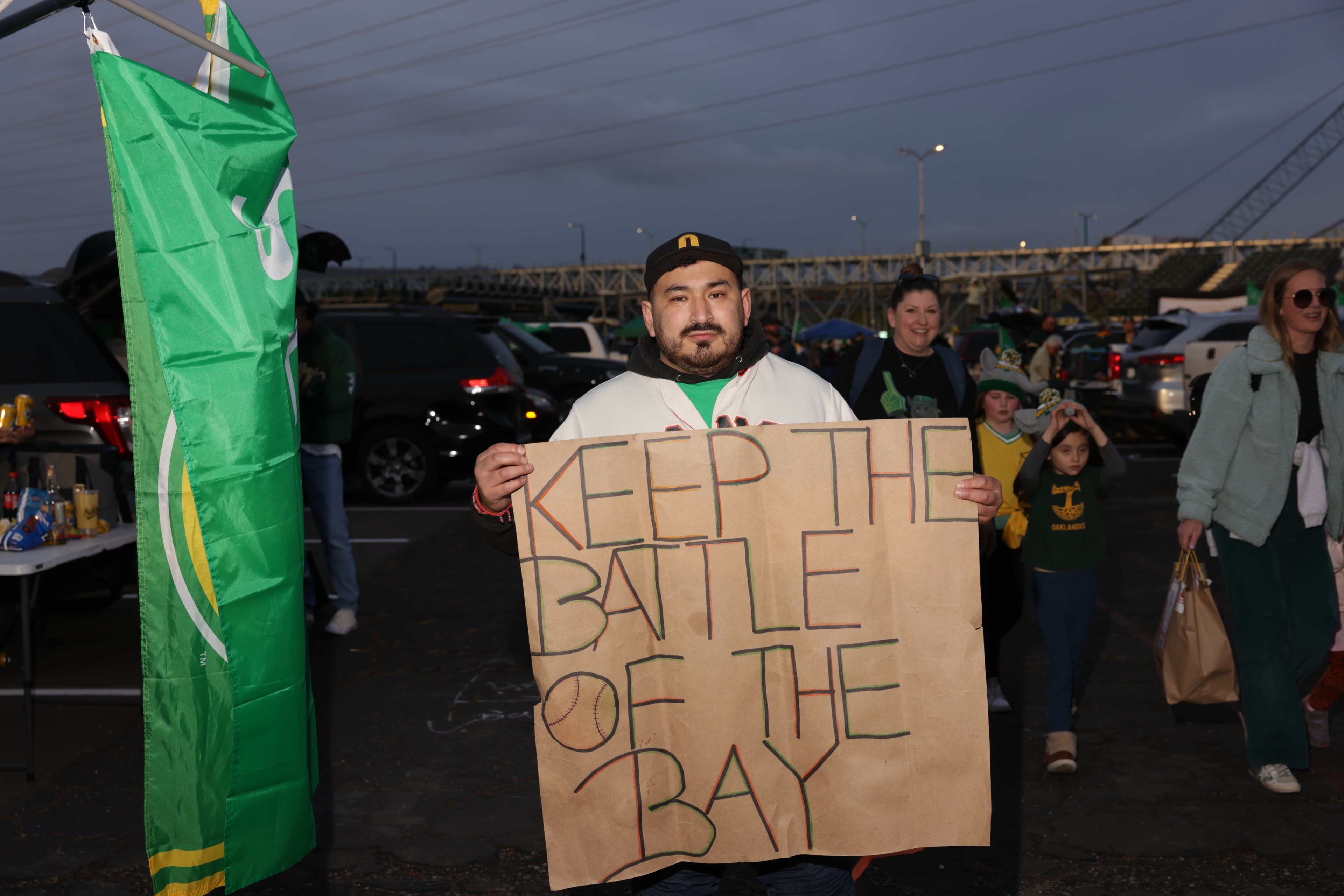 A man holds a sign &quot;KEEP THE BATTLE OF THE BAY&quot; at a sports event tailgate with others in team colors.