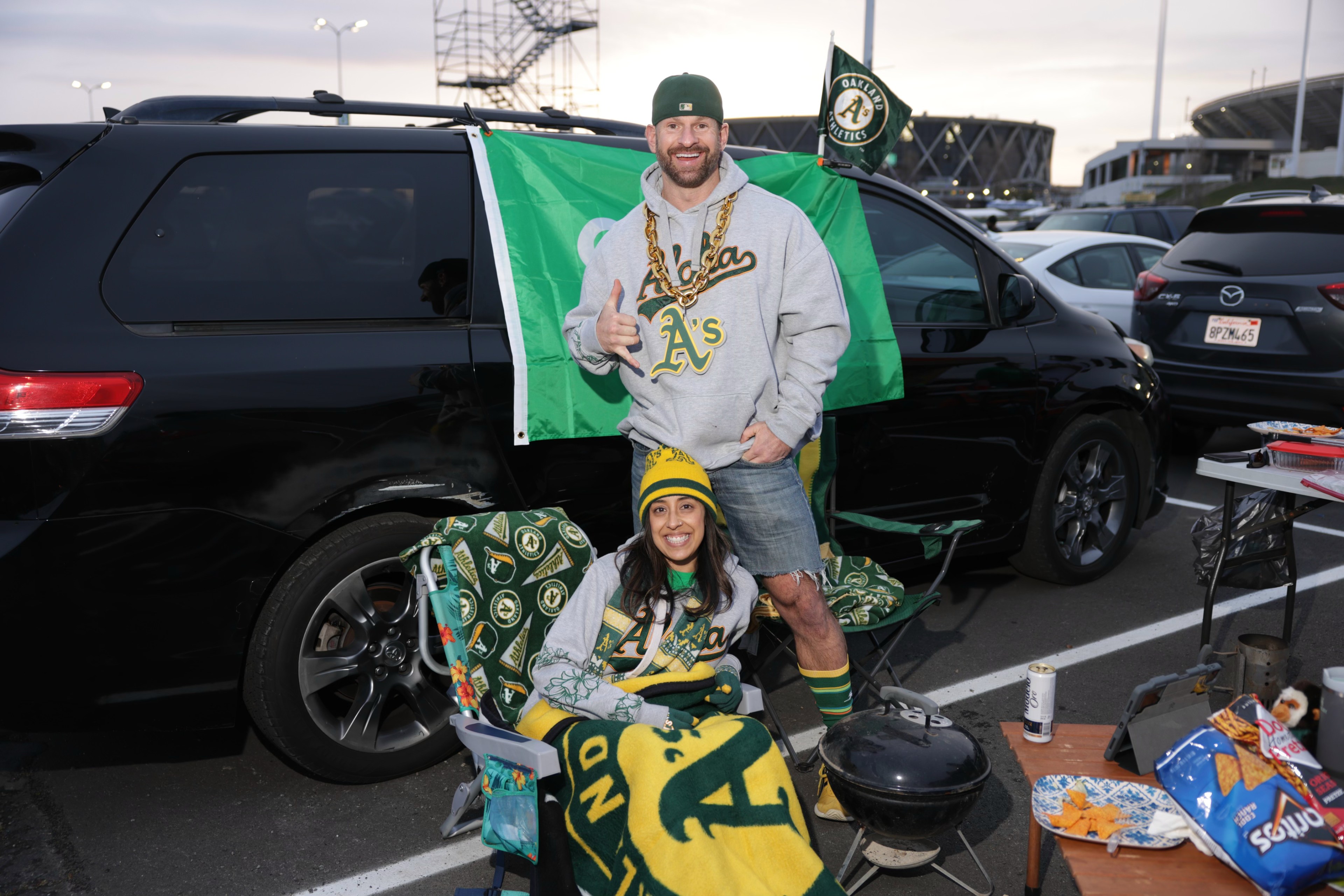 Two fans in Oakland Athletics gear tailgating by their car, with a grill, snacks, and stadium in the background.
