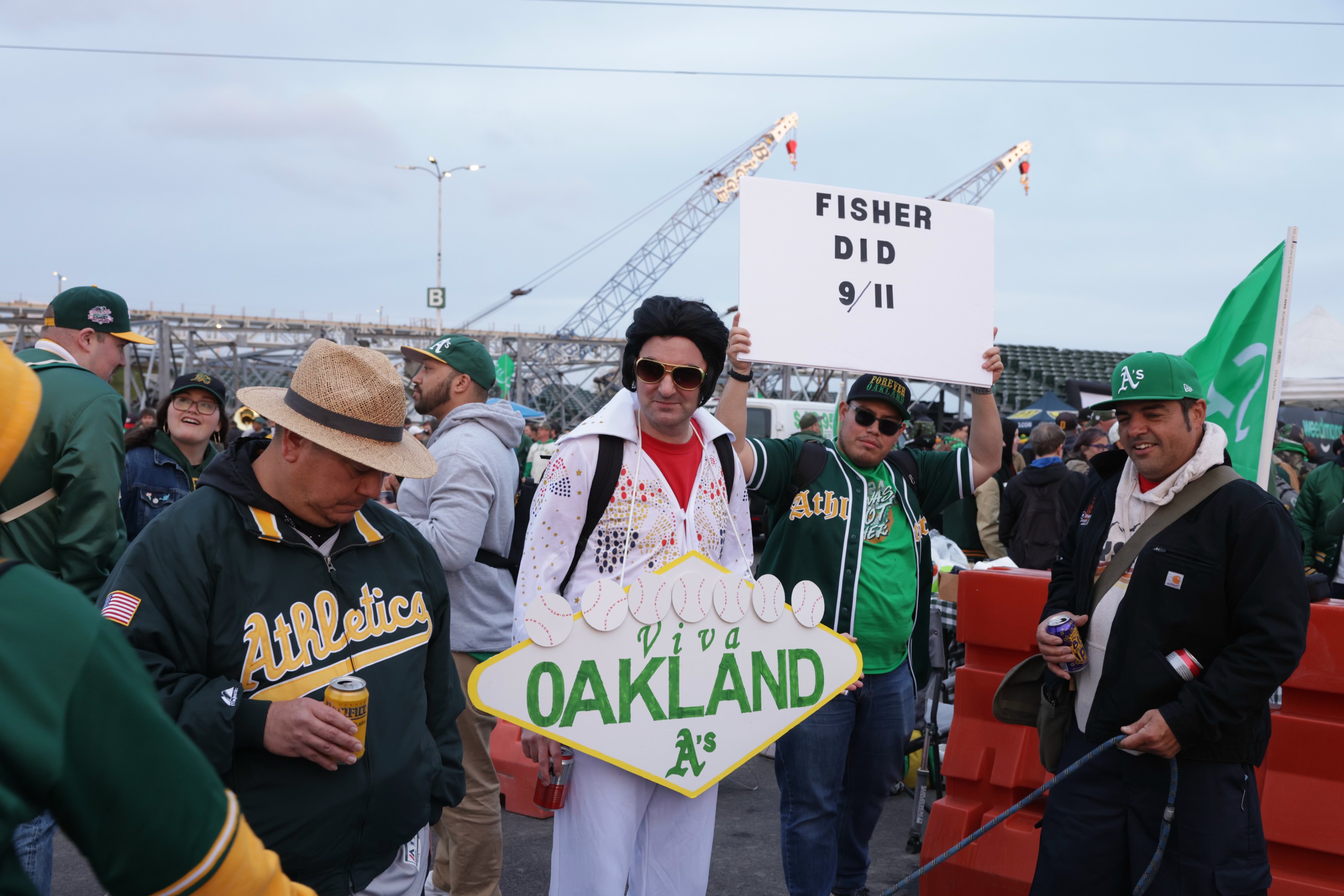 Baseball fans in A's gear, one dressed as Elvis holding a &quot;Viva Oakland&quot; sign, others chatting with drinks, and a bridge in the background.