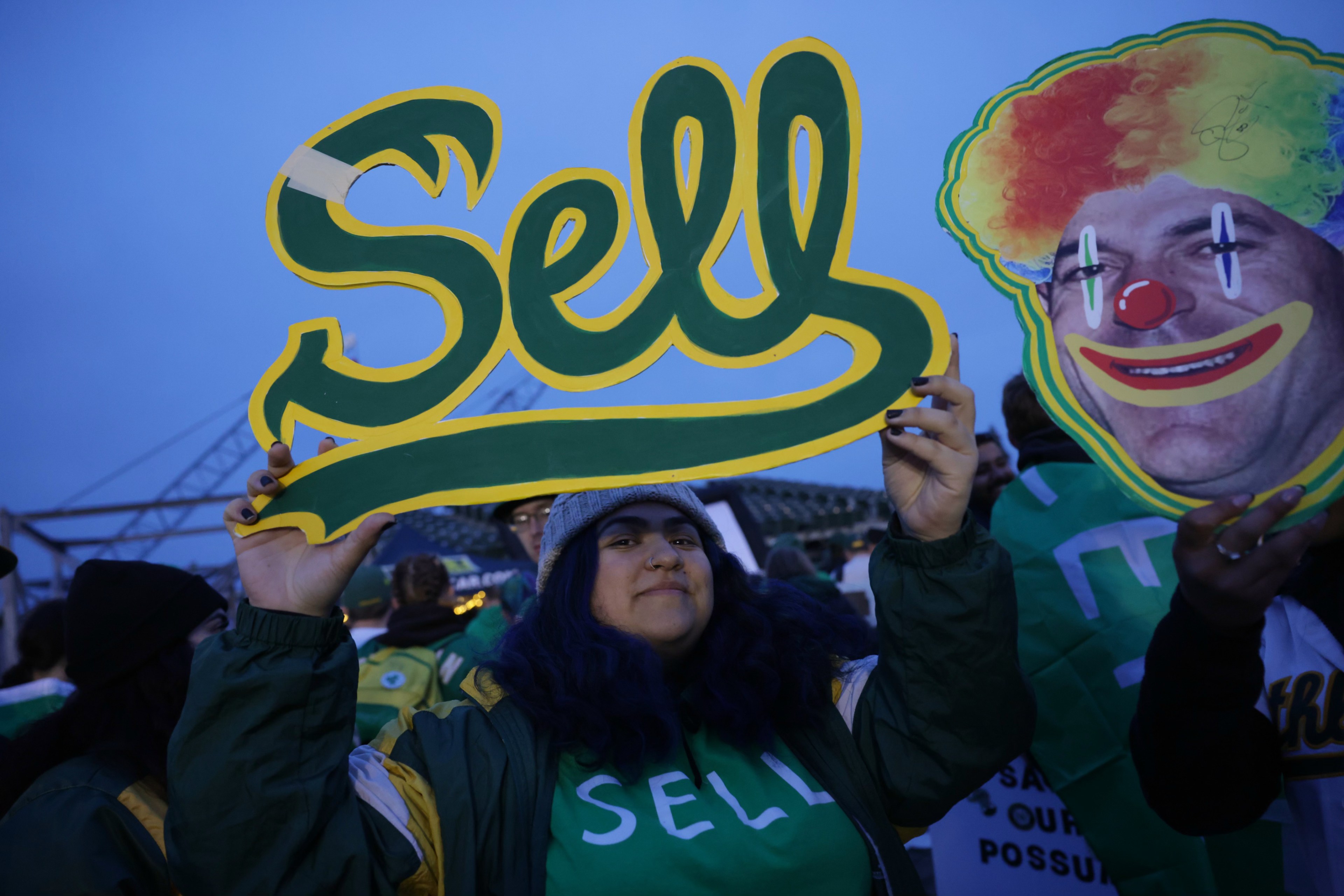 A person is holding up a yellow and green "Sell" sign next to someone with a clown face cutout. They appear to be at an event.