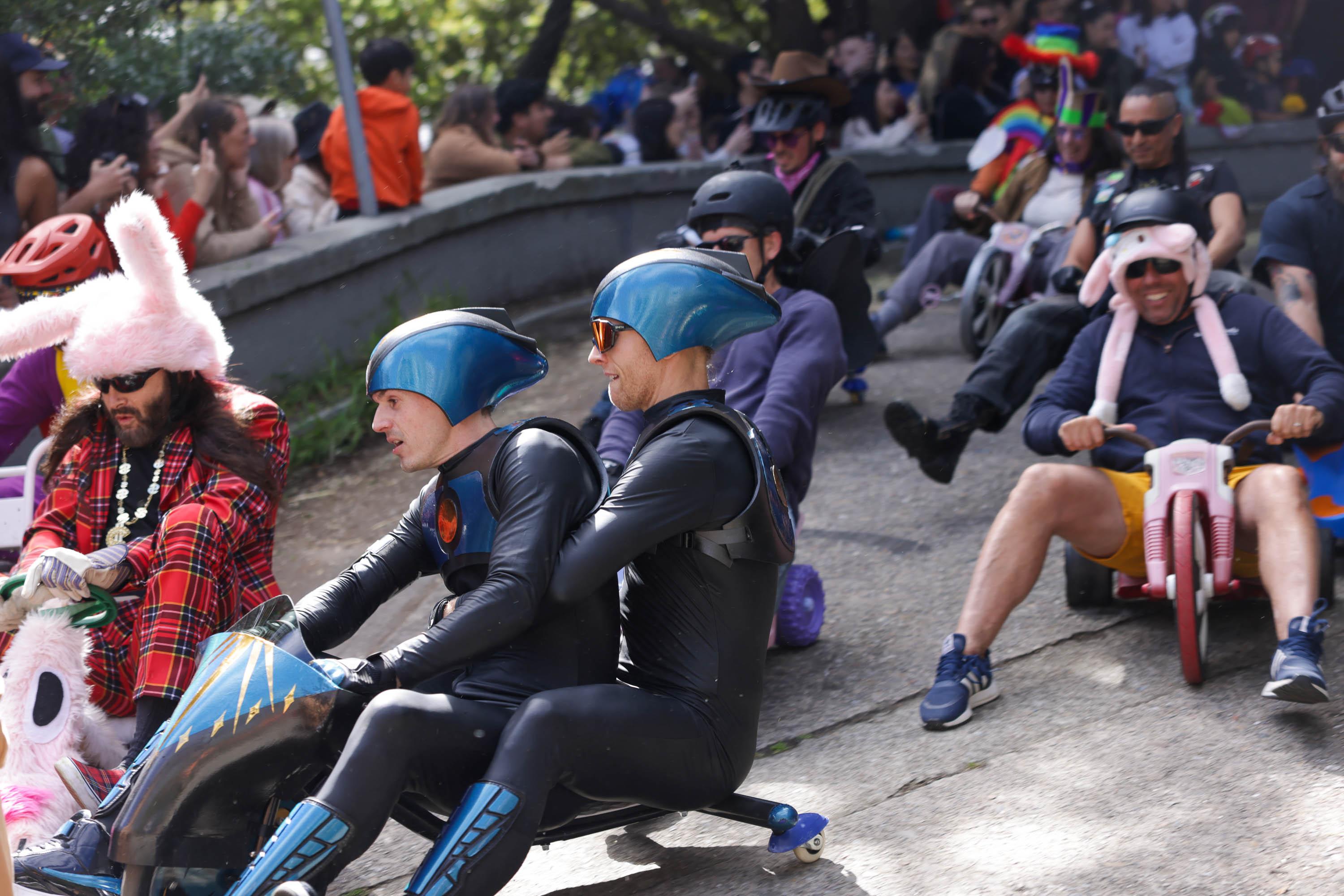 Adults in costumes race downhill on toy bikes; two are dressed as superheroes, others in playful outfits, amidst a watching crowd.