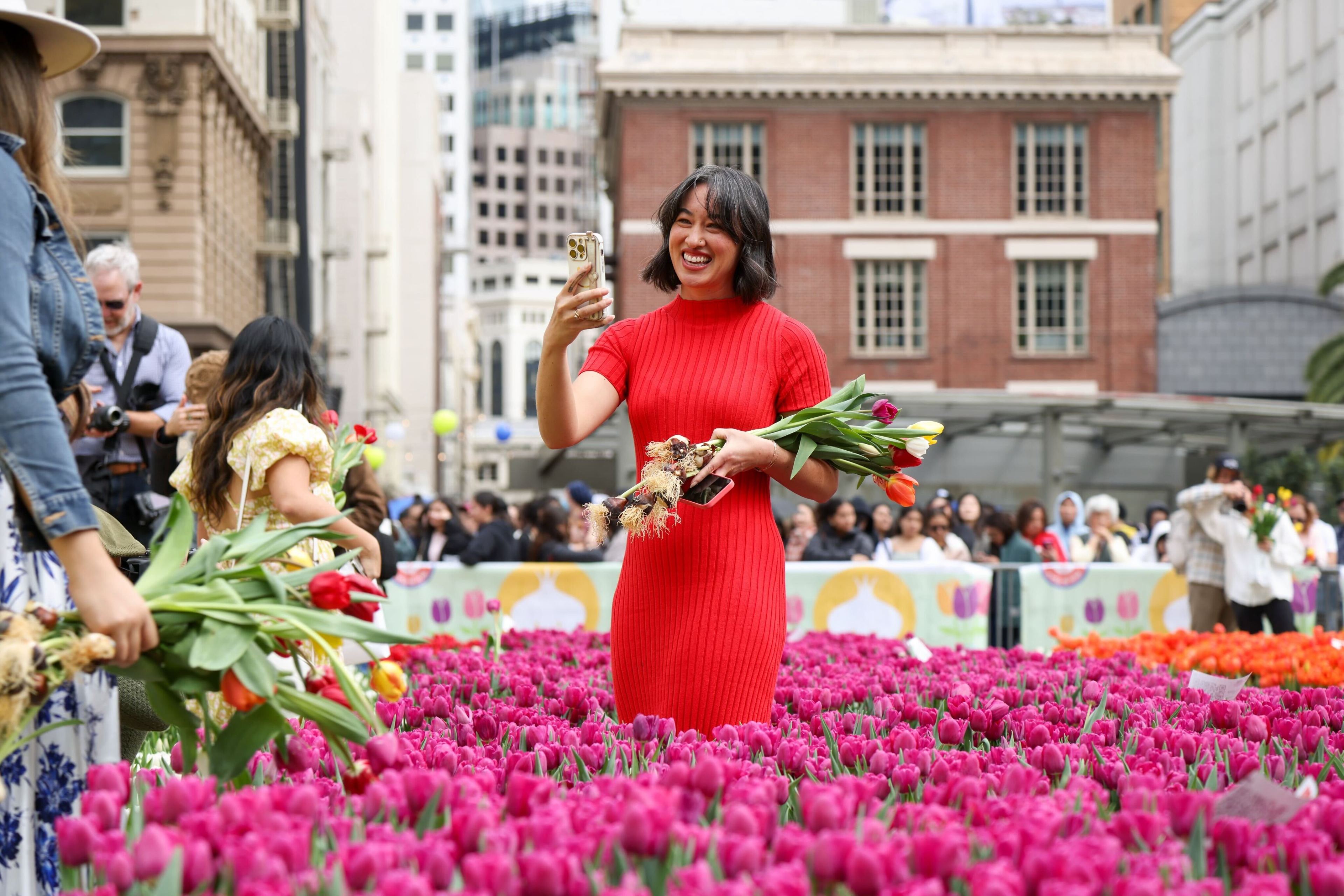 A woman in a red dress smiles among vibrant tulips, taking a selfie with her phone.