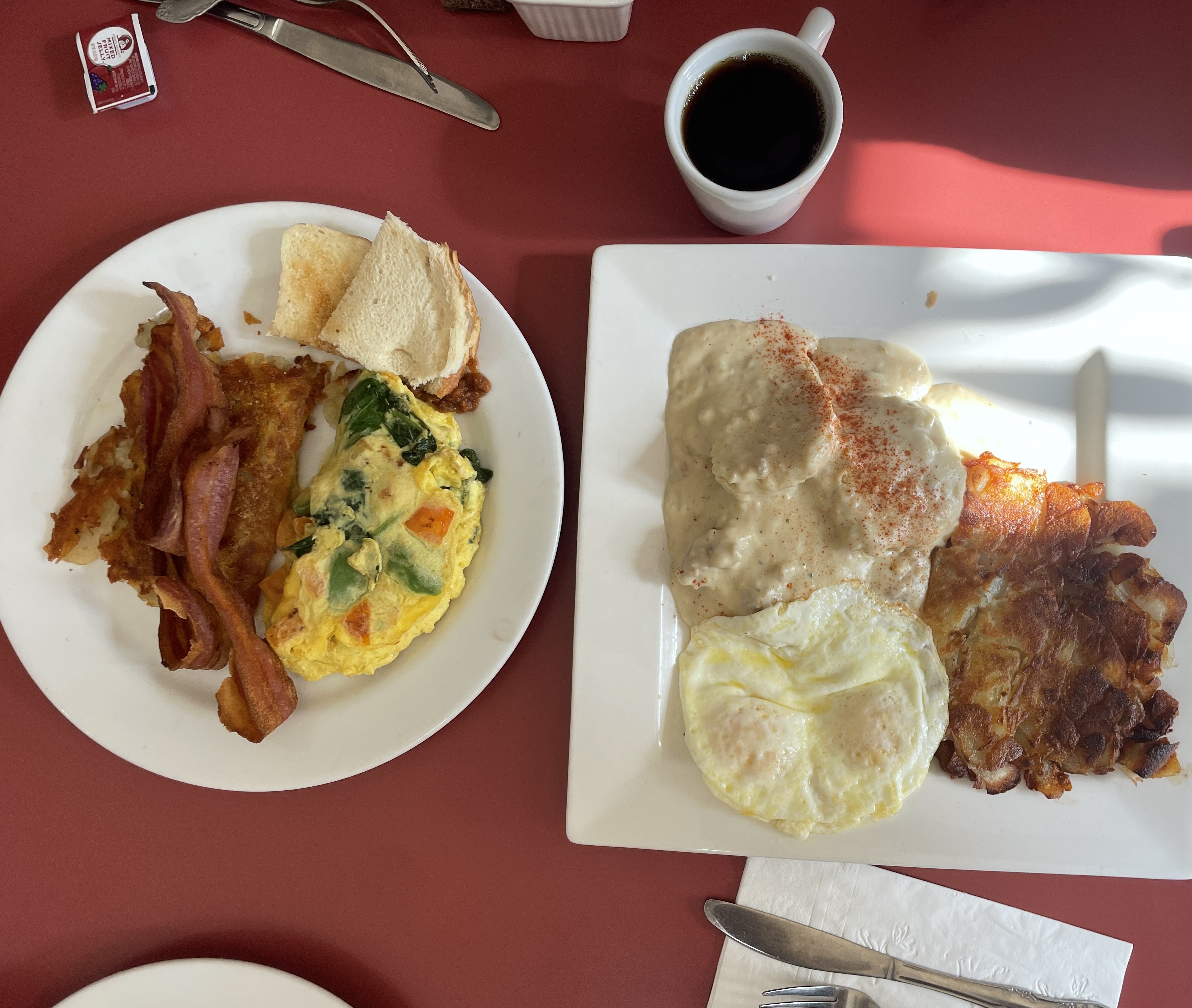 Two breakfast plates on a red table: one with an omelette, bacon, toast; another with eggs, gravy-covered biscuit, hash browns, and coffee.