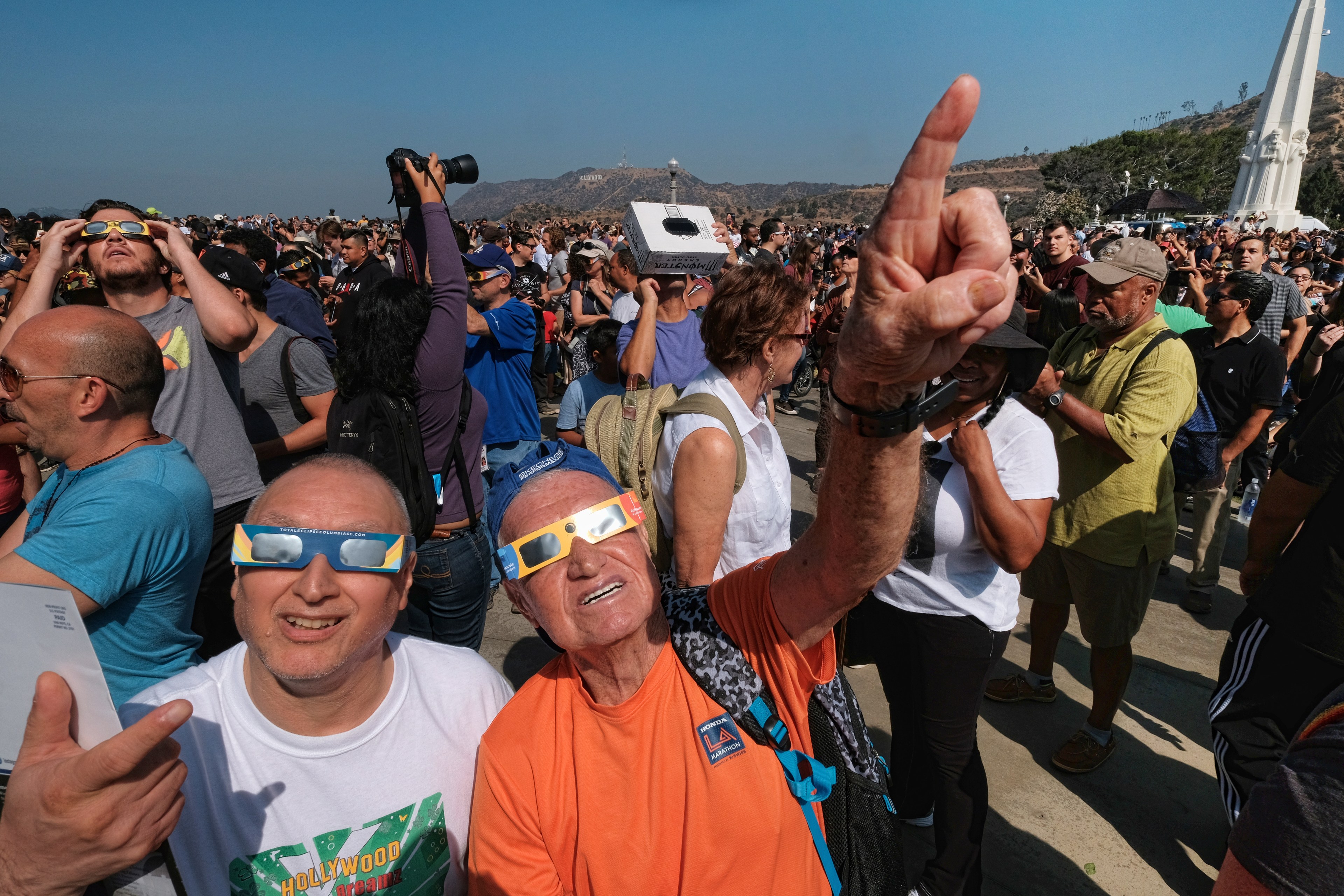 A crowd wearing special glasses looks up at the sky, likely viewing an eclipse, with excited expressions and gestures.