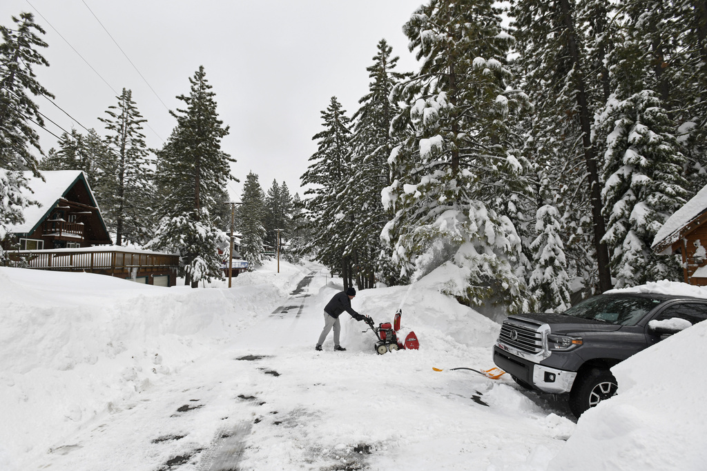 A person clears snow with a red snowblower near a truck; a cabin and snowy trees are in the background.