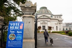 A person walks a dog past a &quot;Vote Here&quot; sign at an ornate building entrance.