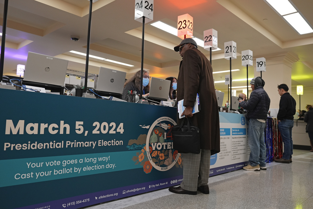 A voter stands at a check-in area with booths and a &quot;March 5, 2024 Presidential Primary Election&quot; sign.
