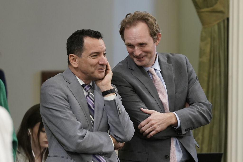Two men in suits are smiling and whispering, conveying a sense of camaraderie or shared secret.