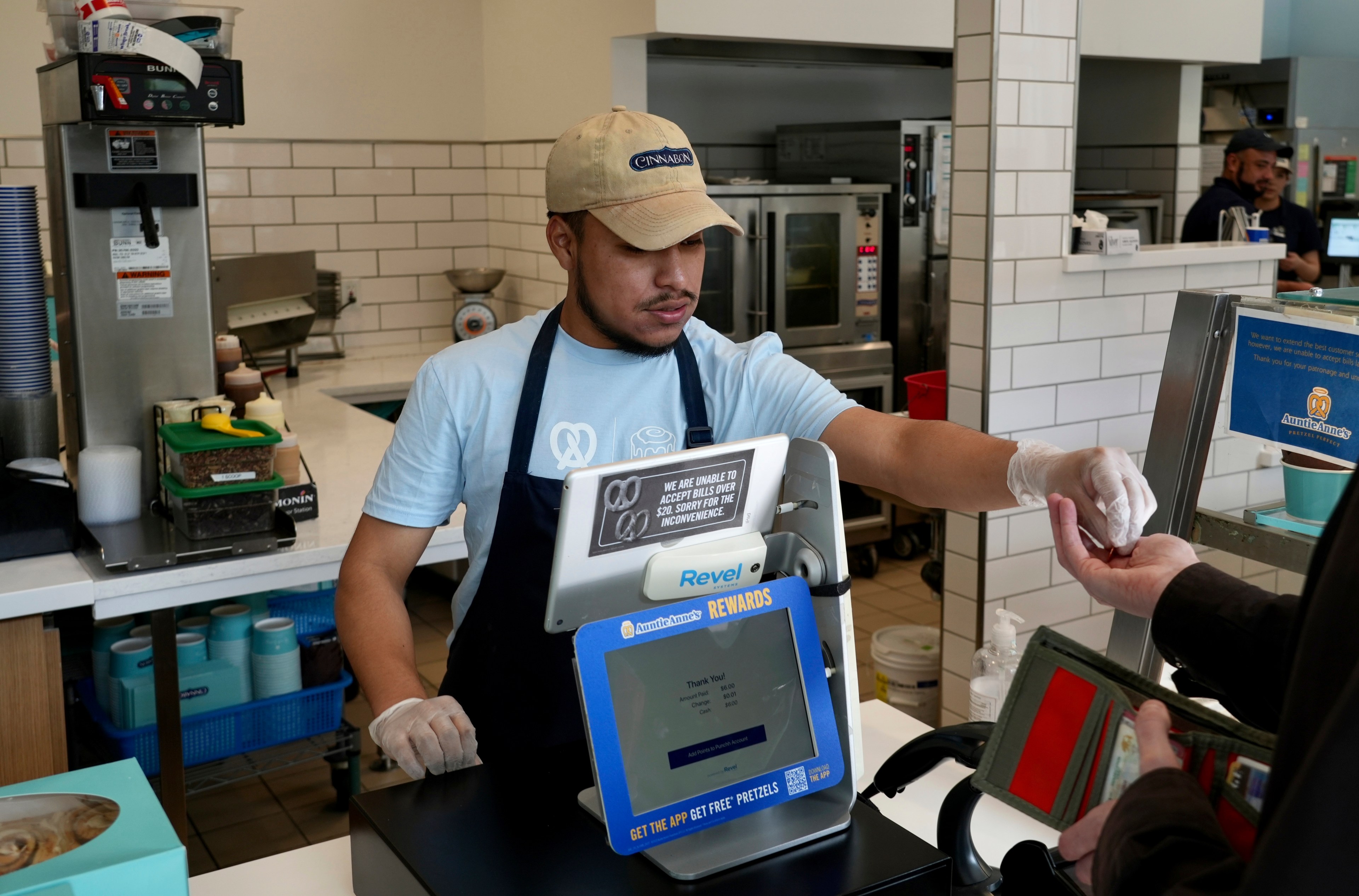 A cashier in a cap and apron hands a receipt to a customer at a fast-food counter, with kitchen equipment in the background.