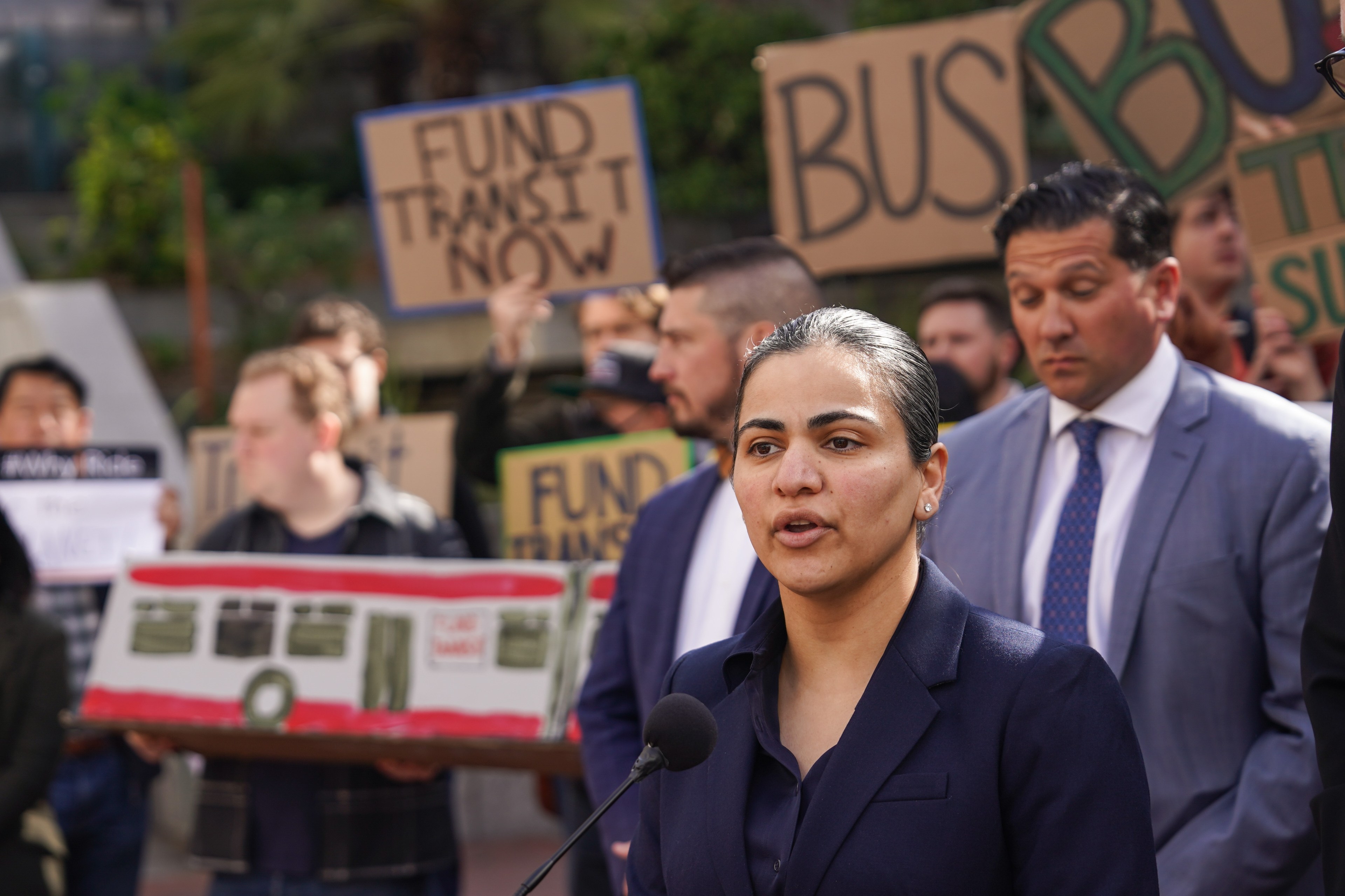 A woman speaks at a rally holding a model bus, with protesters and signs saying &quot;Fund Transit Now&quot; in the background.
