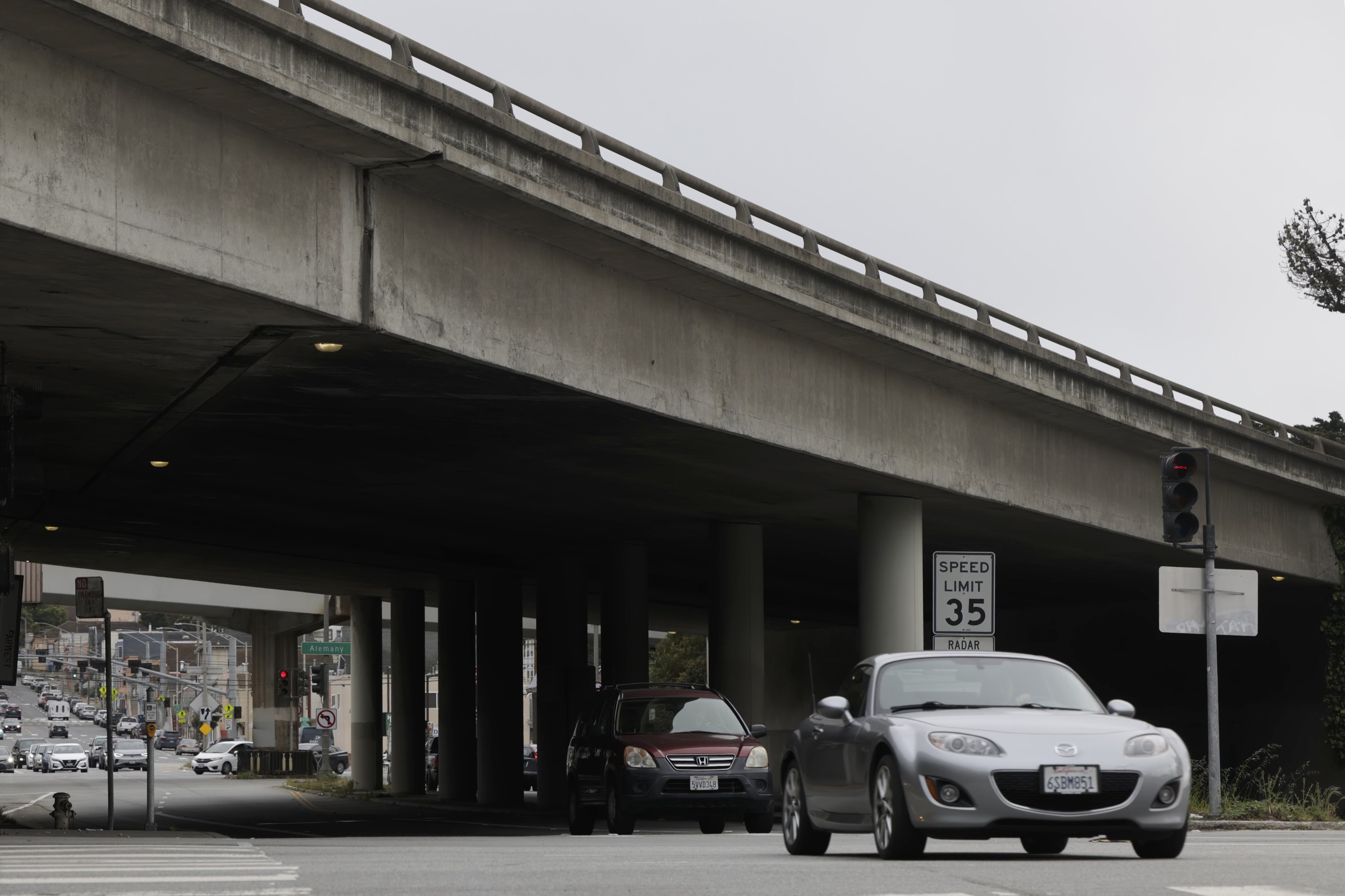 A car waits at a red light beneath an overpass, with a speed limit sign nearby and a busy intersection in the background.