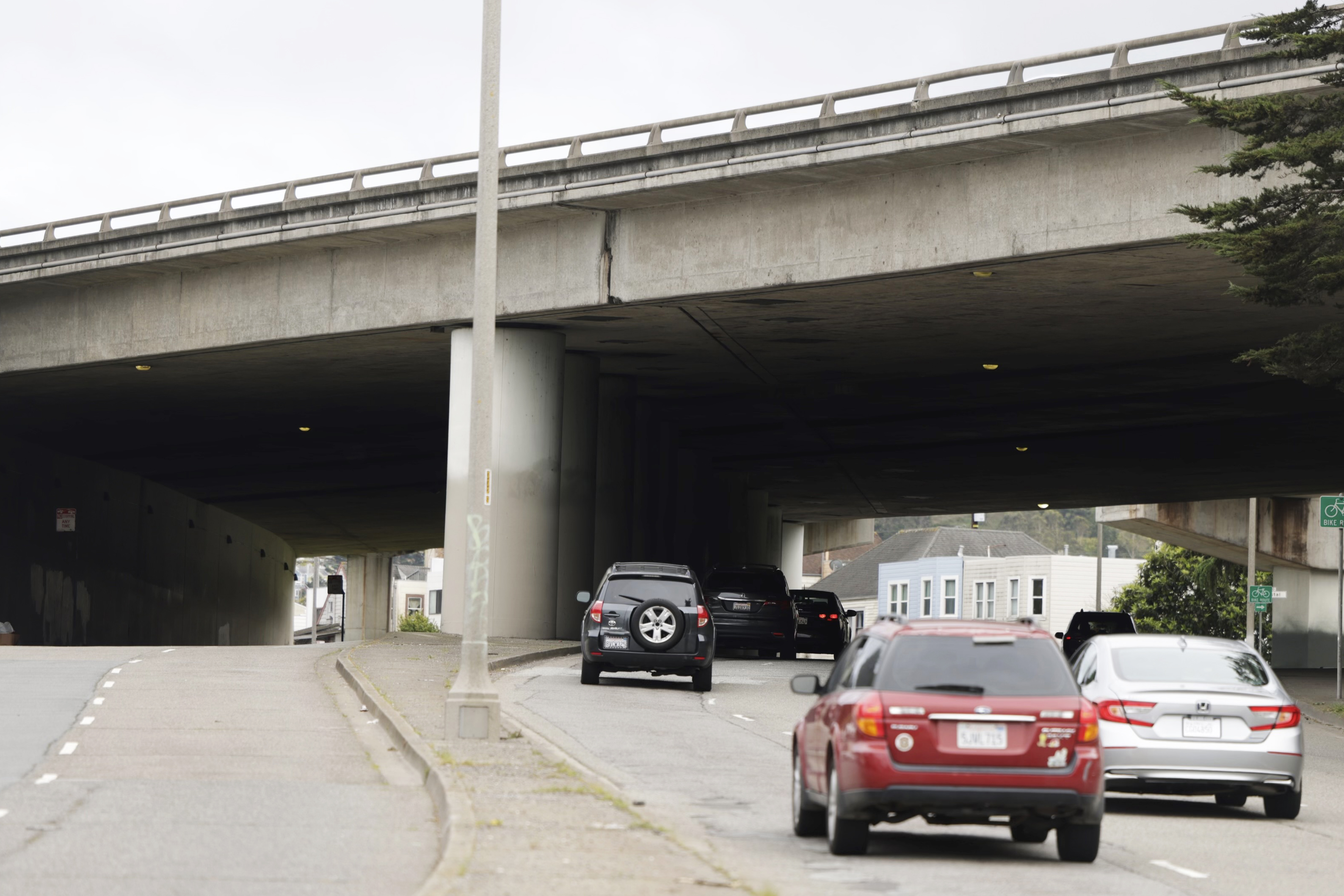 Cars drive under a concrete overpass with small patches of graffiti.