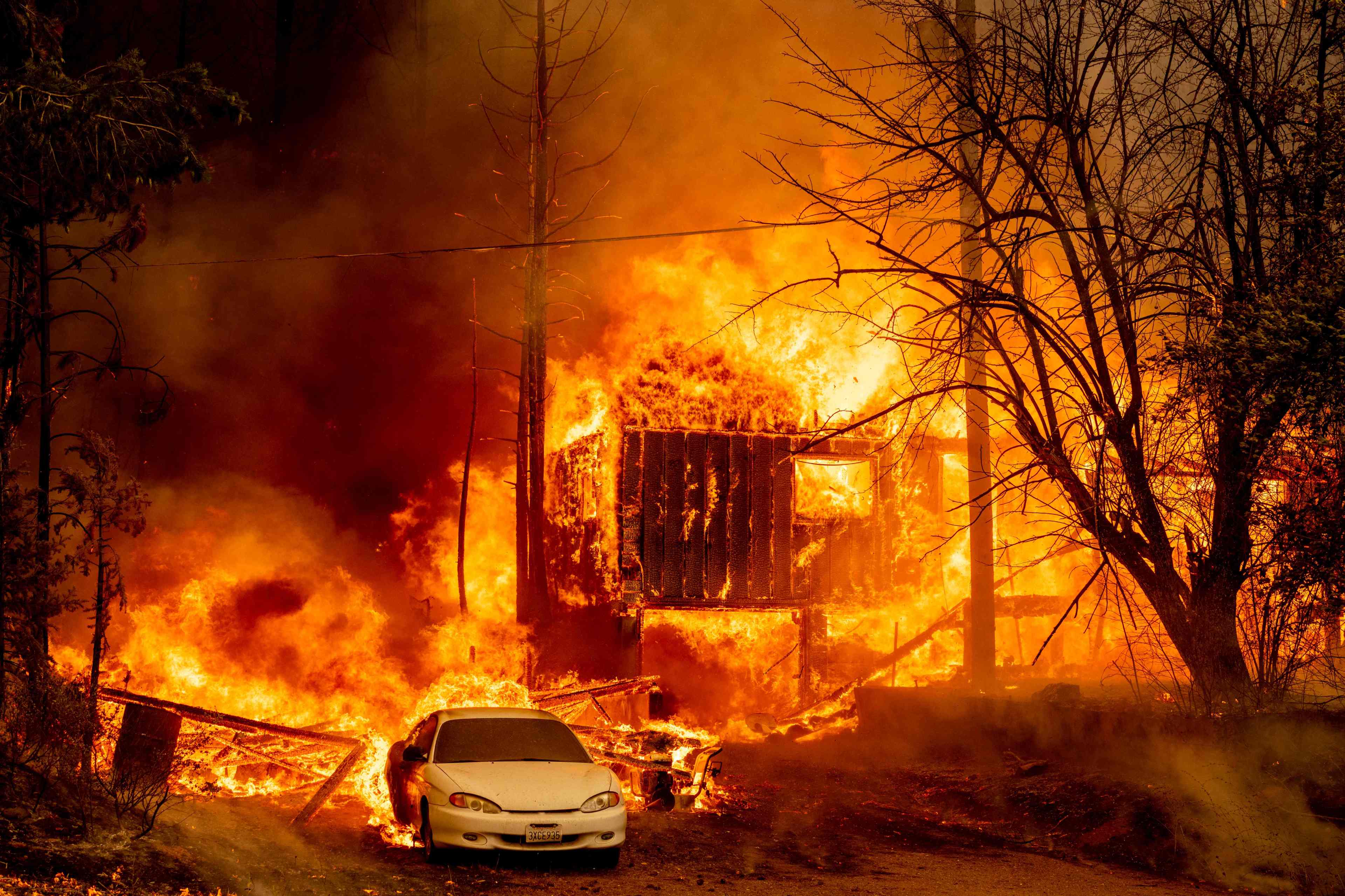 A car parked in front of a house engulfed in flames, with intense fire and smoke around it.
