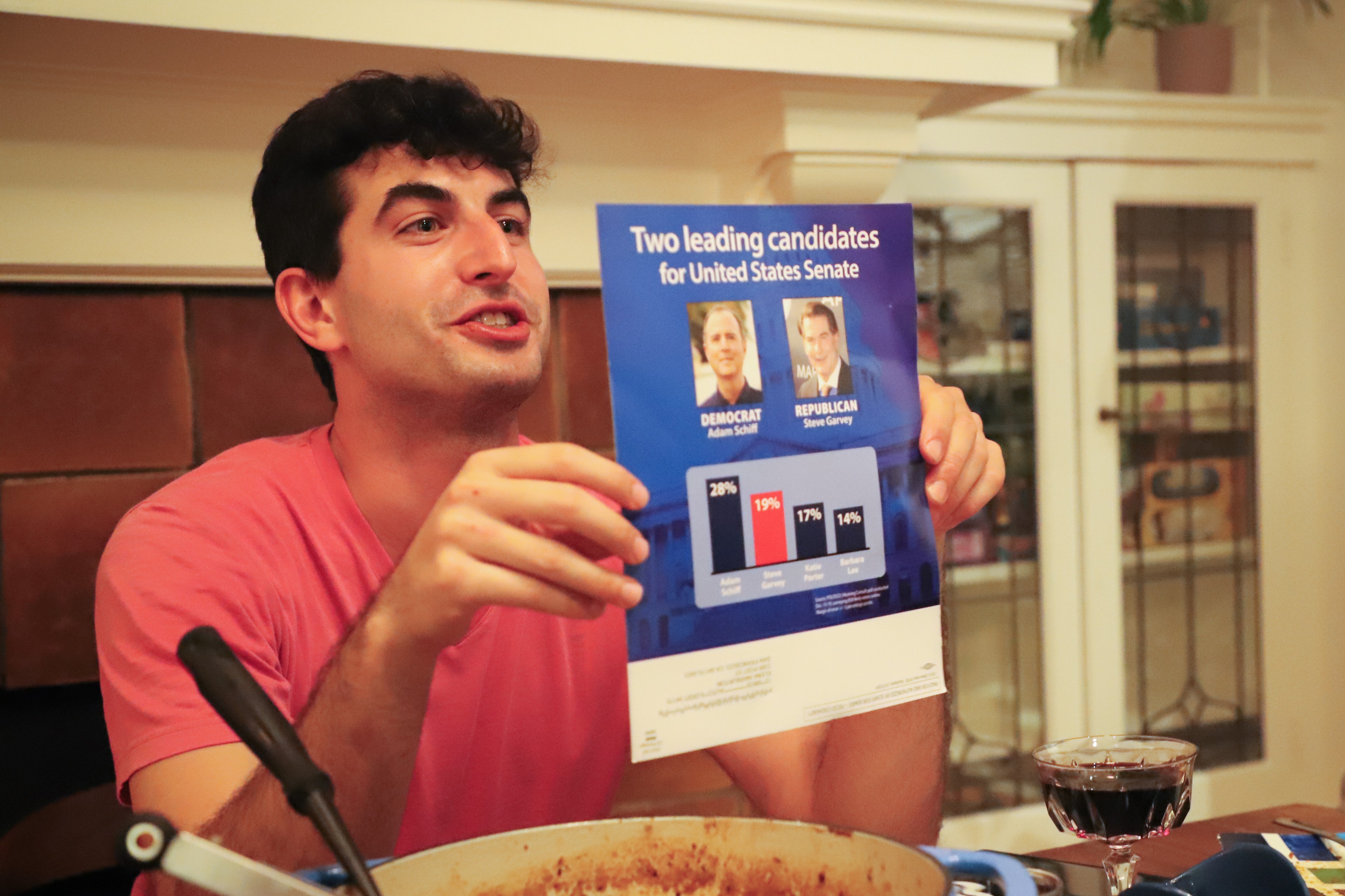 A man holds up a flyer comparing two US Senate candidates at a dinner table with a wine glass nearby.