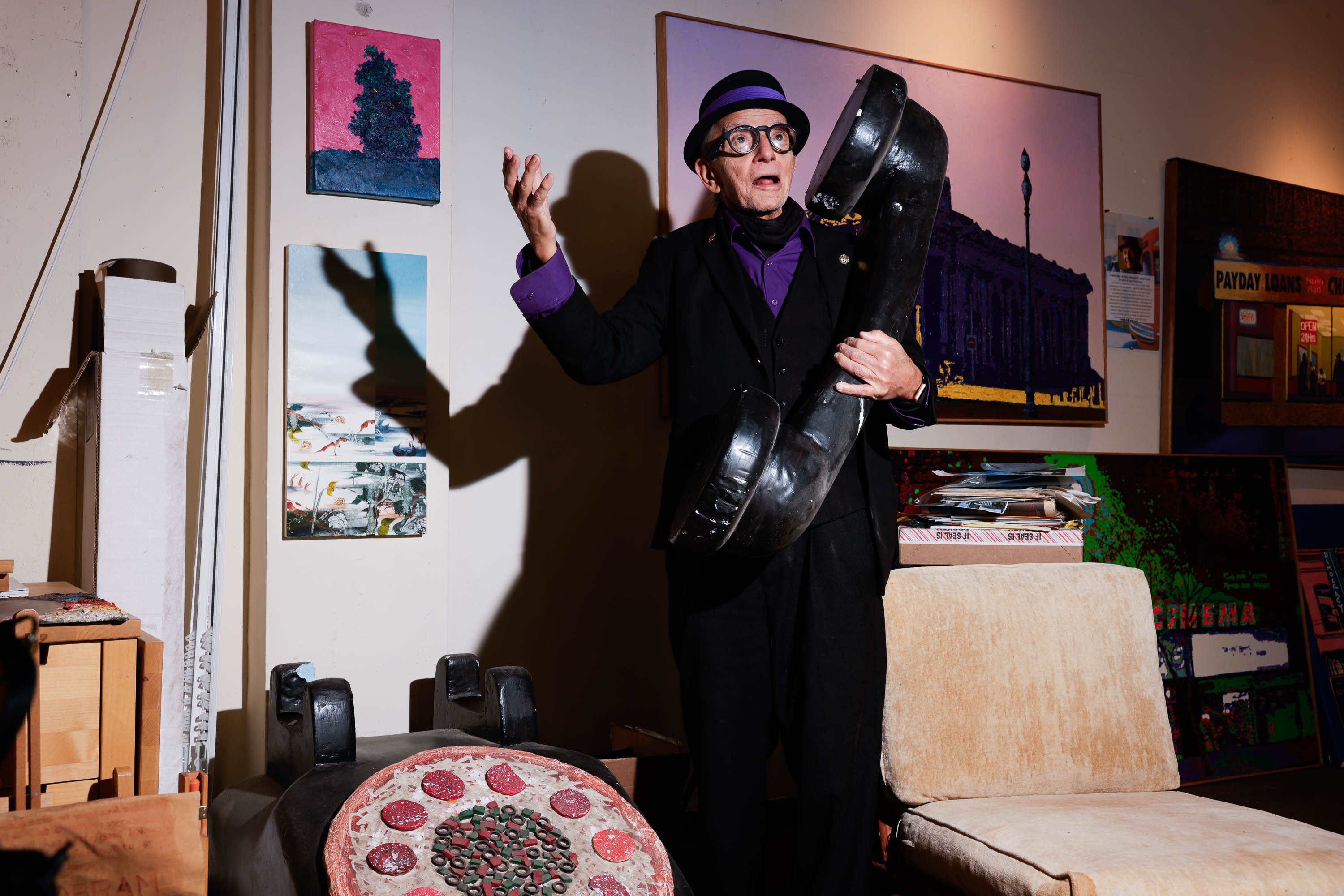 A person in a black suit and hat holds up a large boot, gesturing with other hand, amidst colorful artwork and eclectic decor.