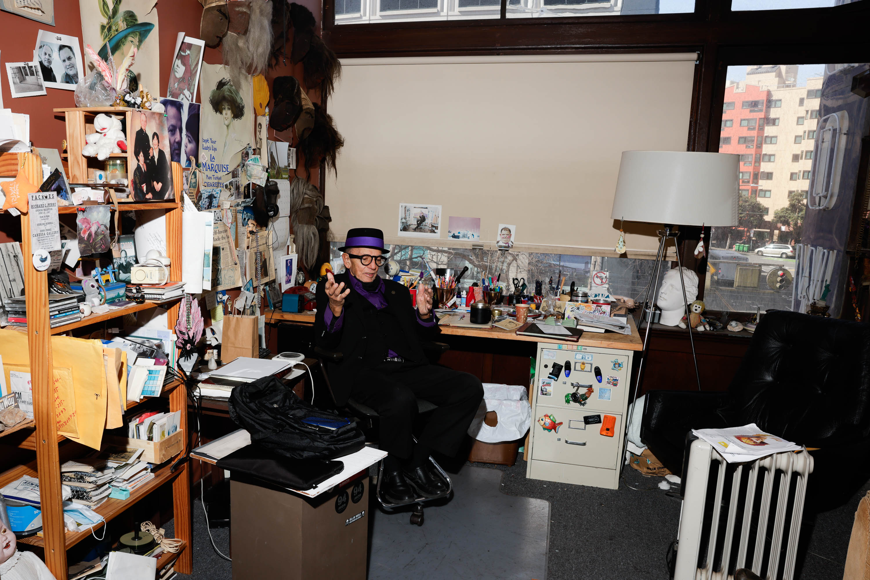 A person sits amidst a cluttered room full of eclectic items, art supplies, and memorabilia.