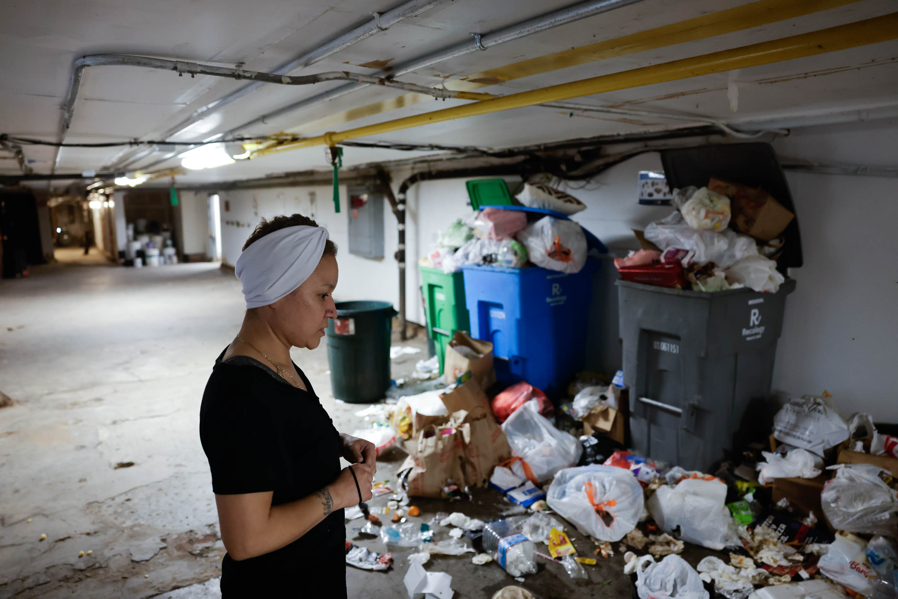 A person stands near overflowing trash bins in a dimly lit garage.