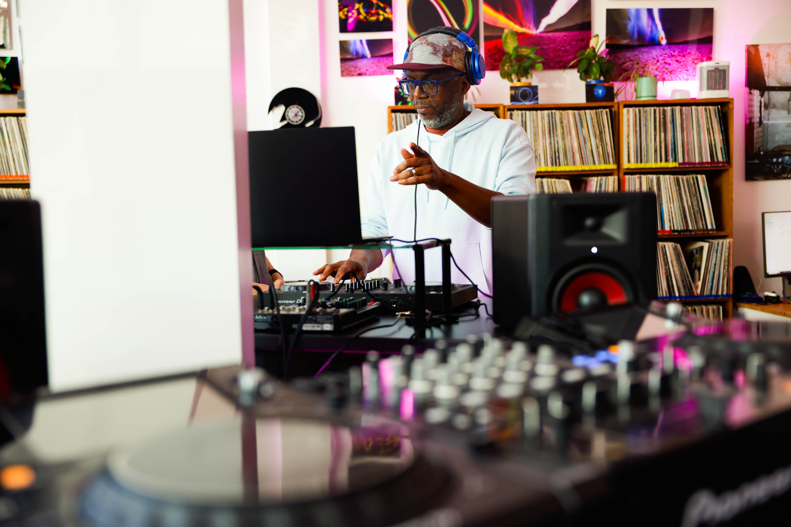 A DJ in a cap and headphones mixes tracks amidst a room full of vinyl records.