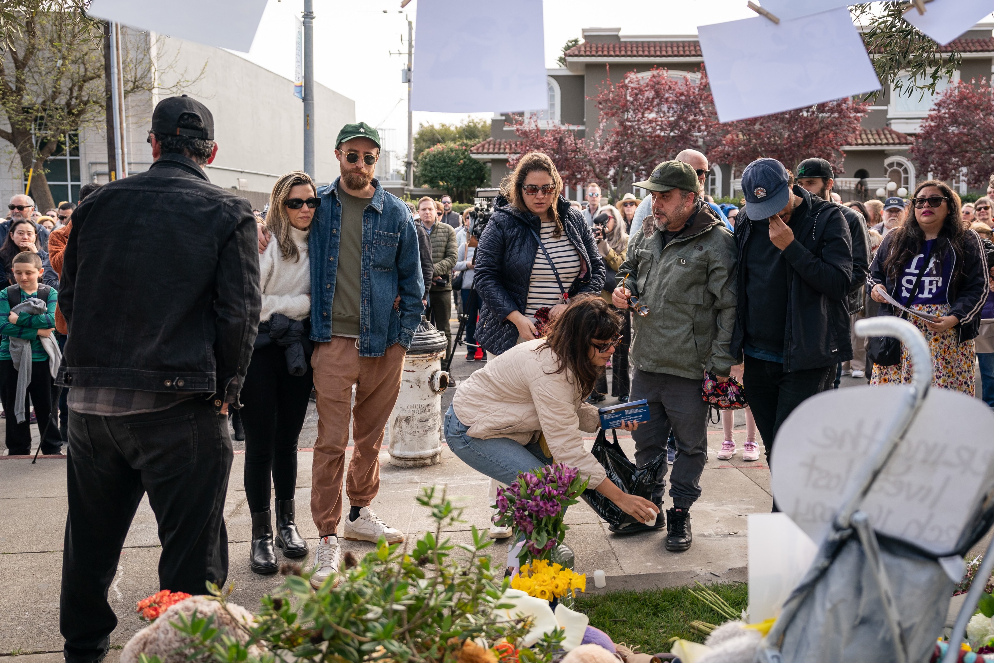 A group of people stands around a street memorial with flowers and messages. Some observe solemnly, while one woman adjusts an object.